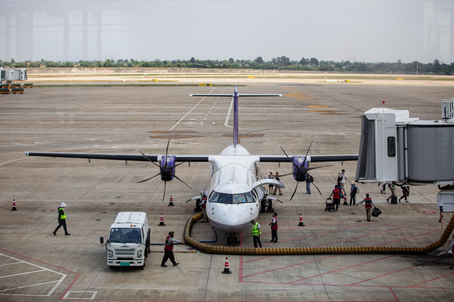 Vista desde el suelo de una pequeña aeronave comercial conectada a una manga de embarque en un aeropuerto en Tailandia, con personal y pasajeros embarcando y desembarcando.