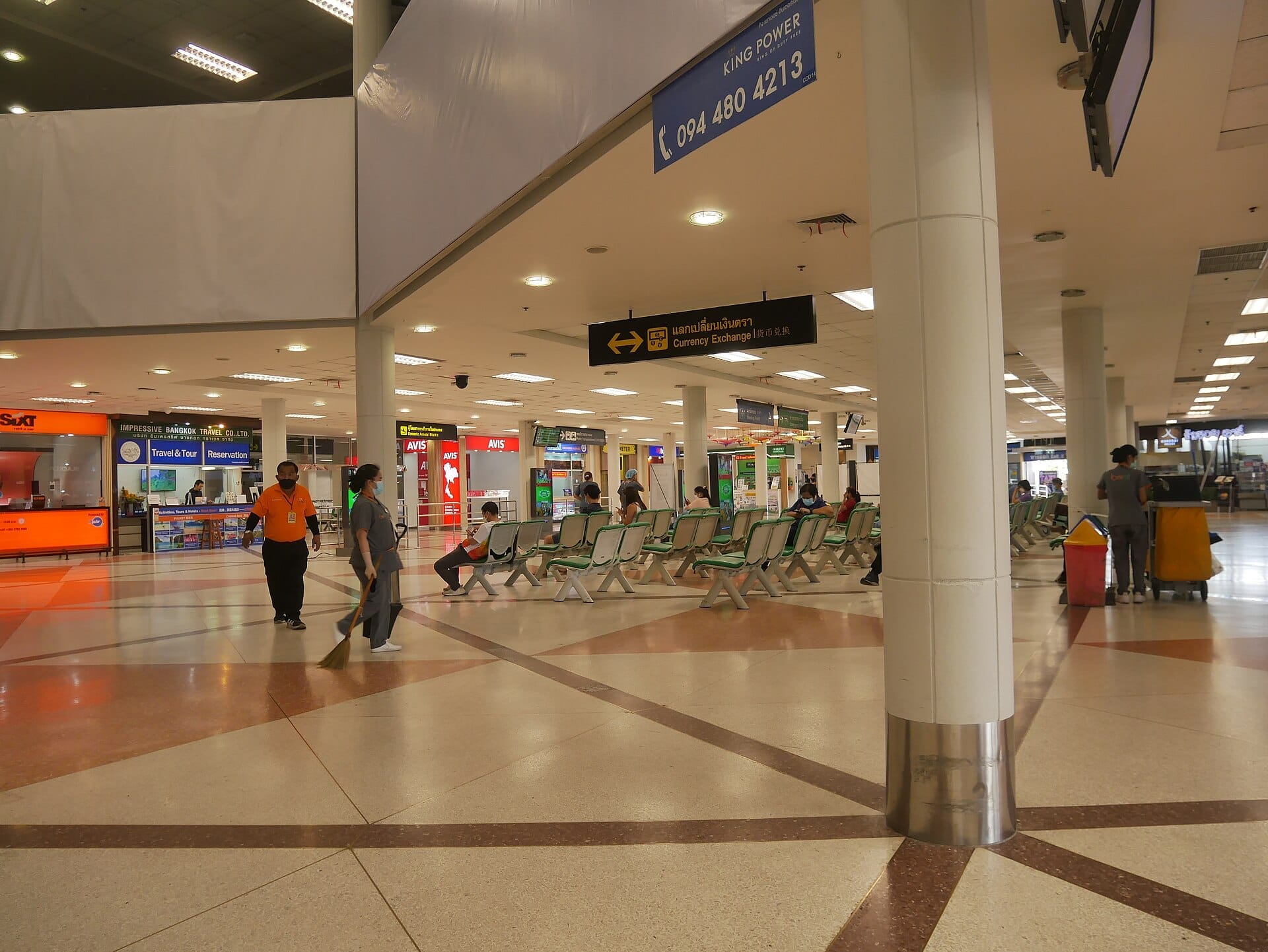 Área de asientos en el Aeropuerto Internacional de Chiang Mai con viajeros, personal de limpieza y letreros de alquiler de autos, cambio de divisas y reservas de tours.