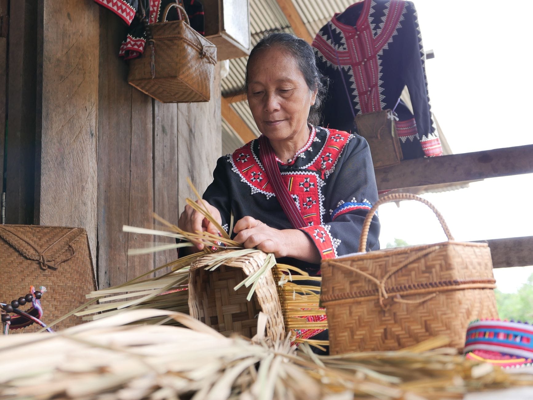 Mujer con traje tradicional tejiendo cestas de bambú a mano en una estructura de madera abierta con artesanías hechas a mano a su alrededor.