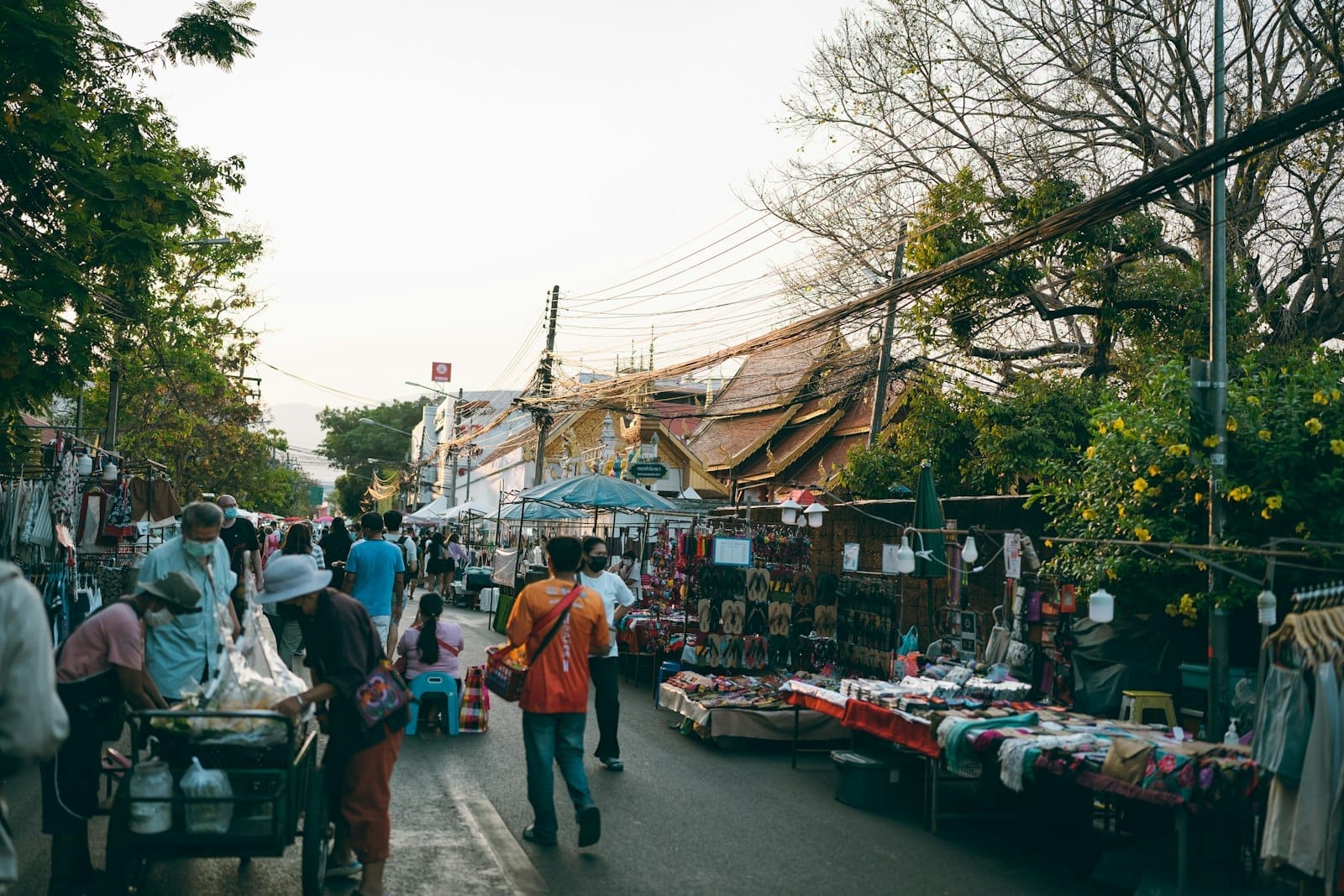 Cảnh chợ ngoài trời nhộn nhịp ở Chiang Mai với người dân đang xem hàng tại các gian hàng và mái chùa hiện ra ở phía sau.