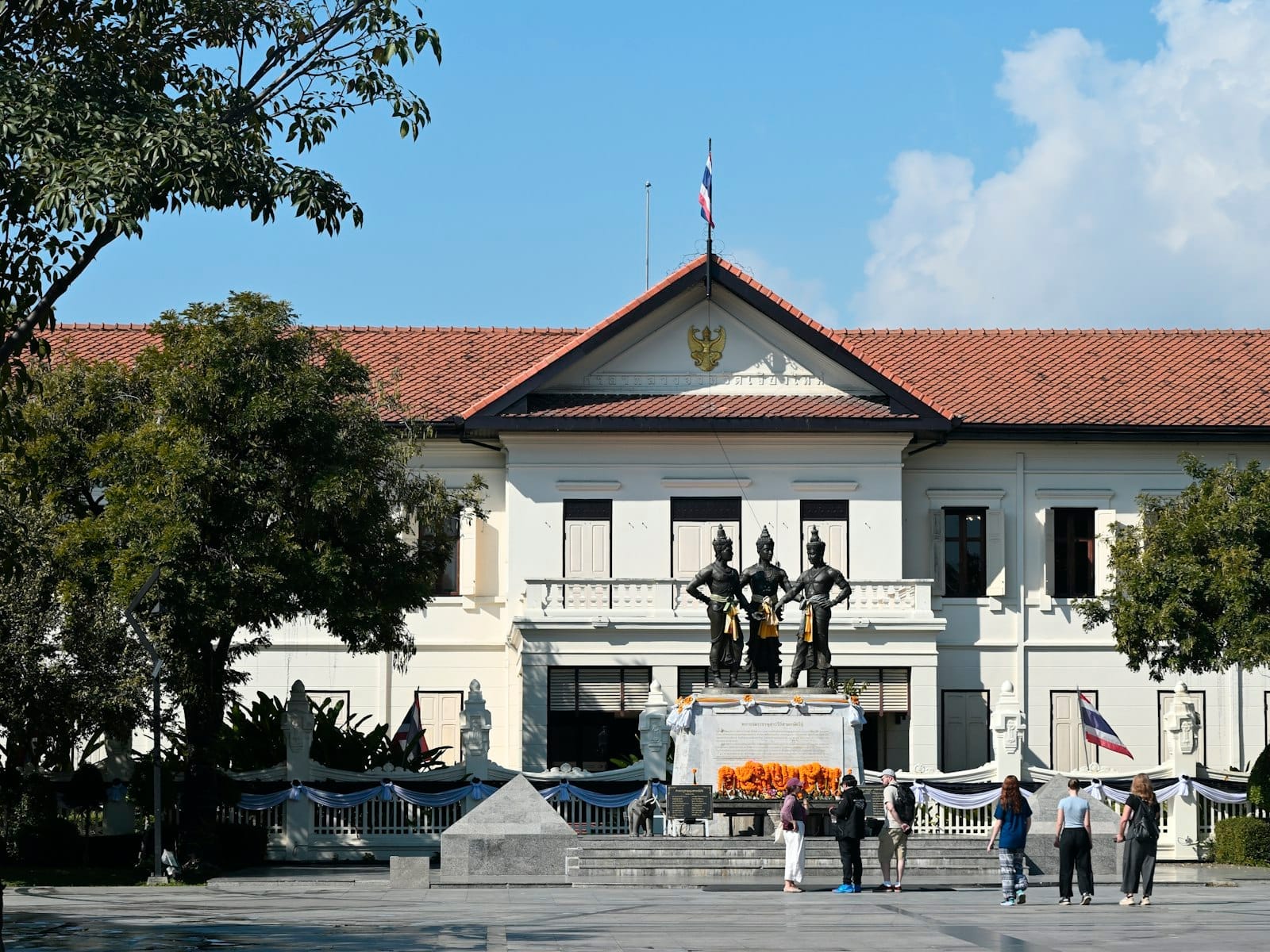 Mặt tiền phong cách thuộc địa của Chiang Mai City Arts and Cultural Centre với Tượng đài Ba Vị Vua, được bao quanh bởi cây cối và du khách trong một ngày nắng đẹp.