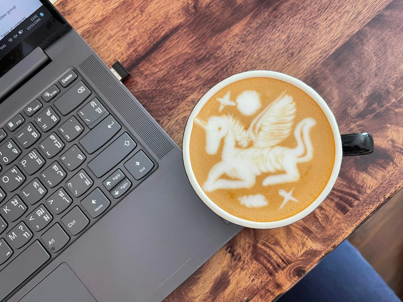 Ly latte với hình nghệ thuật kỳ lân bên cạnh laptop trên bàn gỗ, gợi lên không gian làm việc ấm cúng tại quán cà phê.