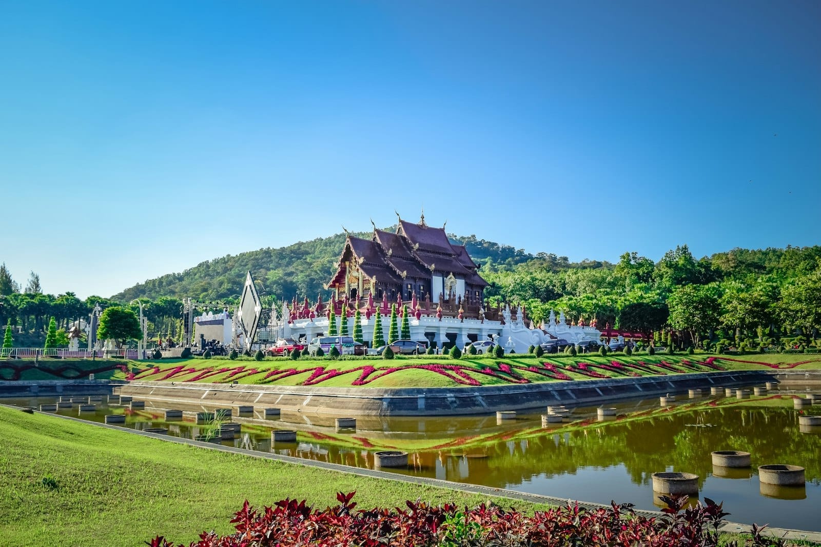 Pabellón de templo de estilo Lanna ornamentado rodeado de jardines y estanque reflectante en el Royal Park Rajapruek, Chiang Mai, bajo un cielo azul despejado.