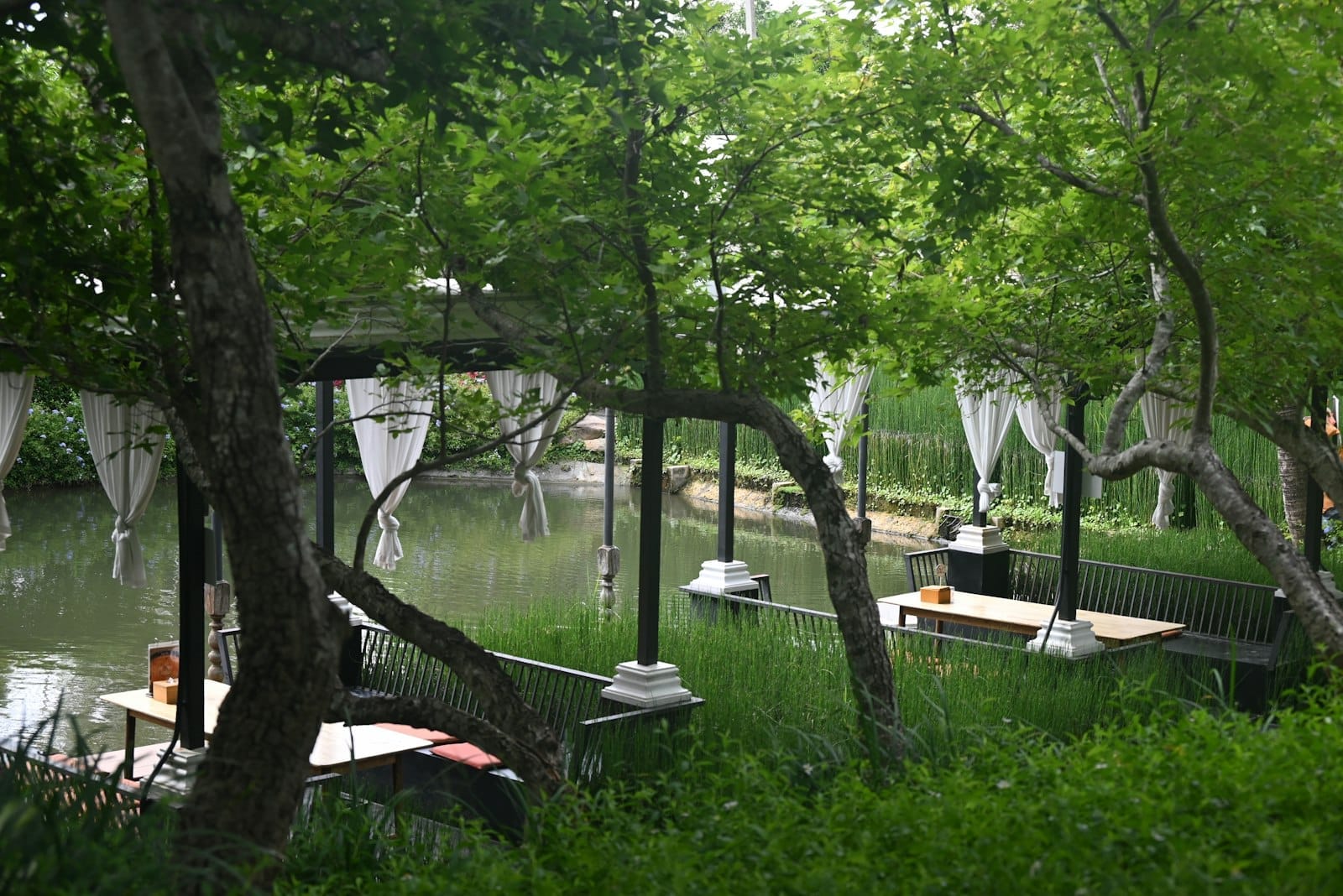Mesas de comedor al aire libre junto al río bajo doseles blancos, rodeadas de exuberante vegetación y agua, creando una atmósfera romántica y tranquila.