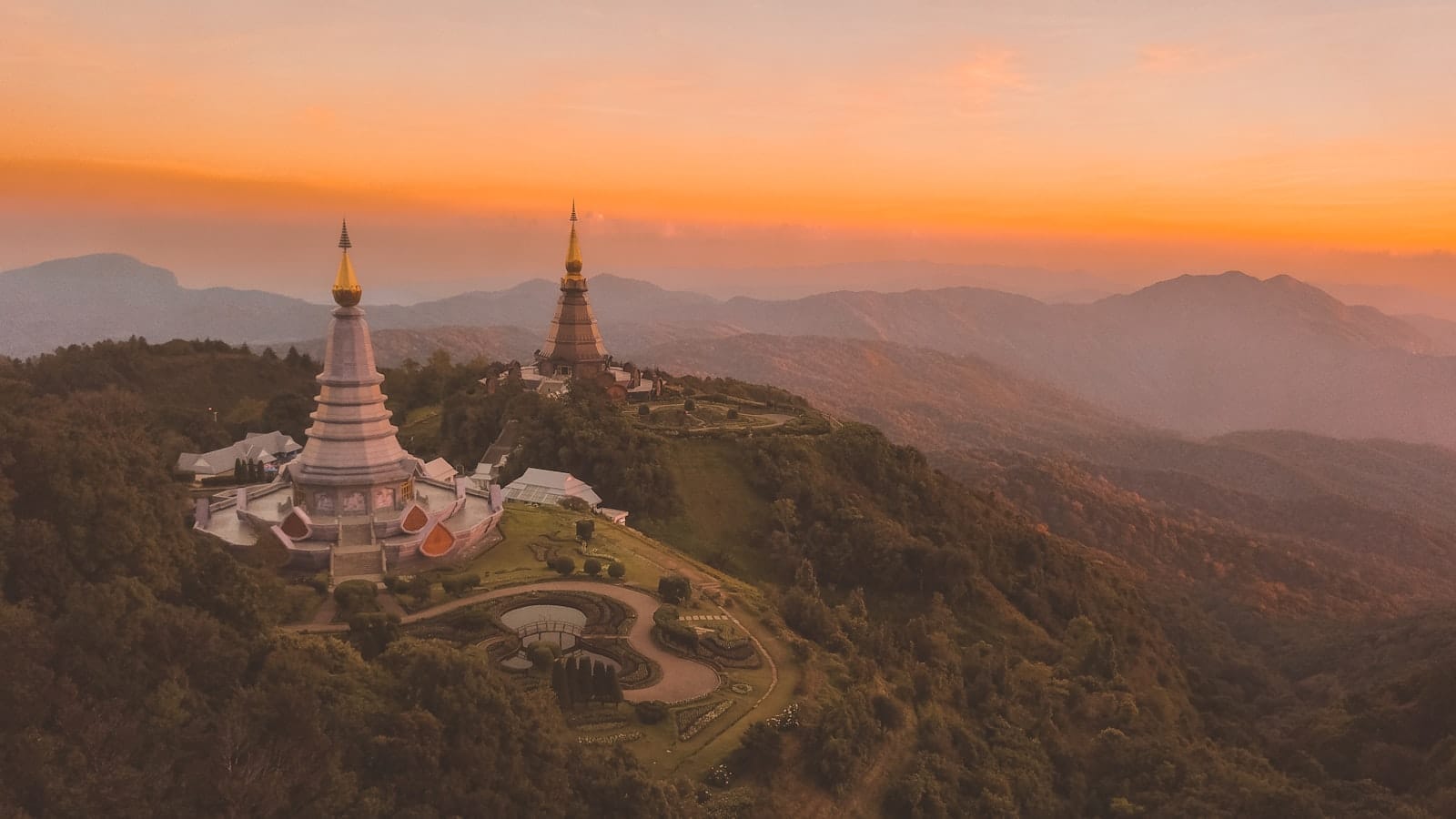 Dos pagodas icónicas en una exuberante montaña verde al atardecer en Chiang Mai, con un cálido cielo anaranjado y colinas en la distancia, creando una atmósfera romántica y tranquila.