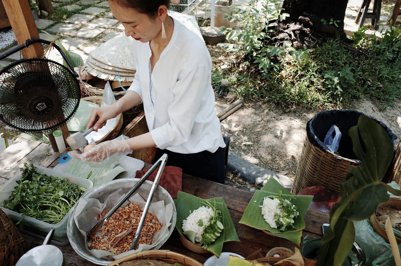 Mujer preparando ingredientes en una estación de cocina al aire libre rodeada de hierbas frescas y verduras en un entorno de jardín.
