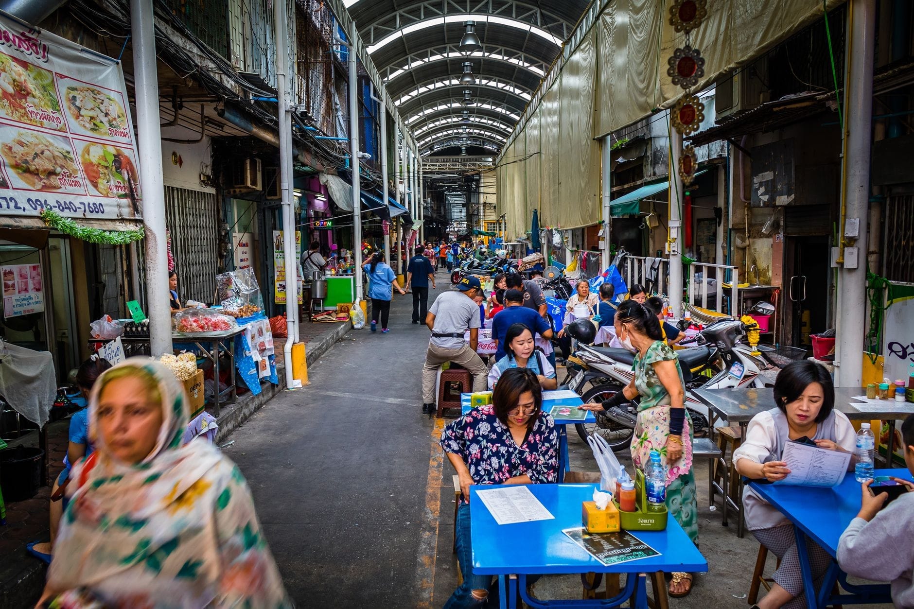 Con hẻm chợ địa phương nhộn nhịp ở Chiang Mai với người dân dùng bữa tại các bàn xanh, người bán hàng và mái che phía trên.