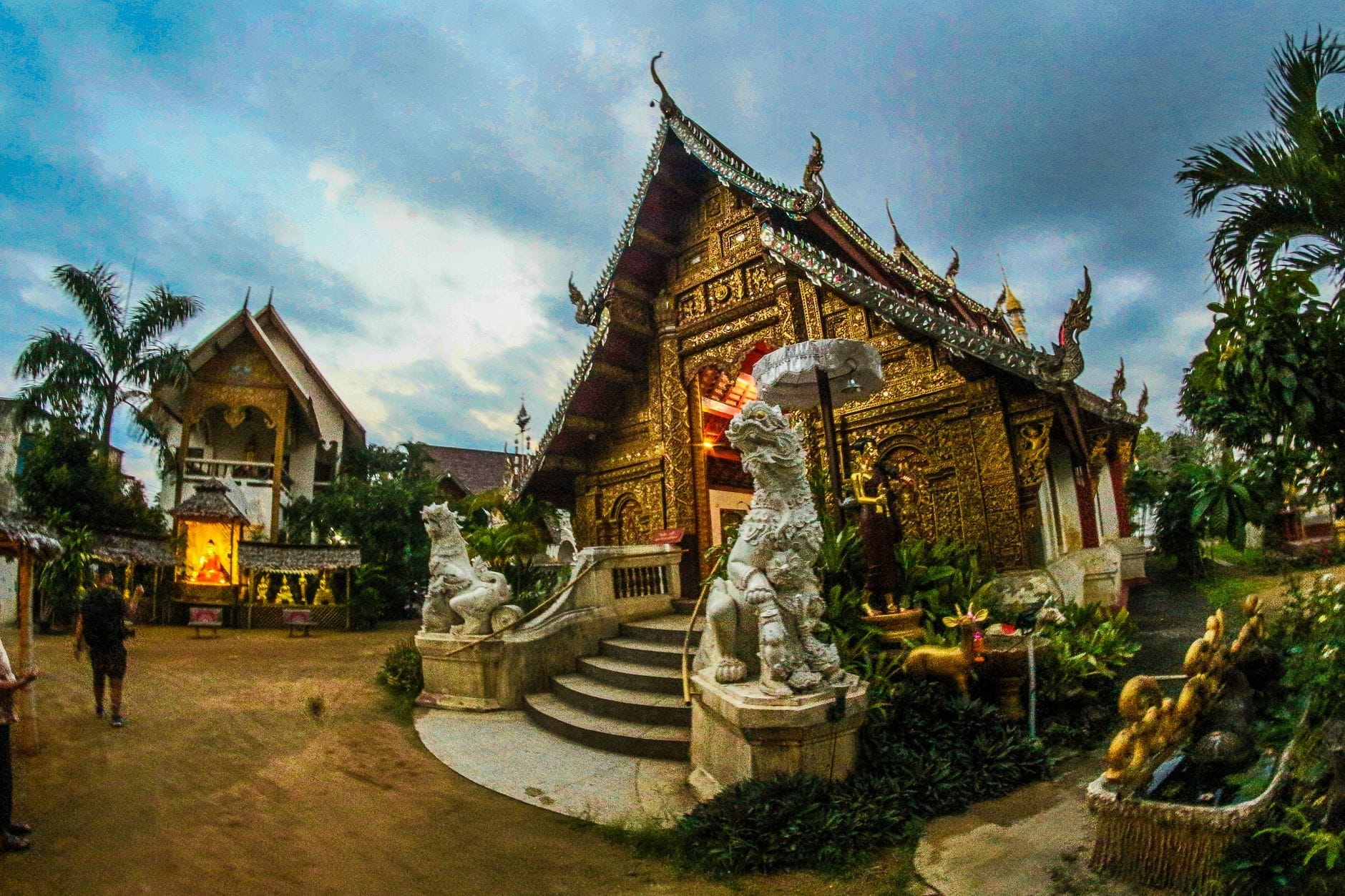 Ngôi chùa nhỏ được trang trí cầu kỳ ở Chiang Mai với các chi tiết mạ vàng và tượng thần hộ vệ, bao quanh bởi cây cối xanh tươi trong khung cảnh yên tĩnh và đầy huyền bí.