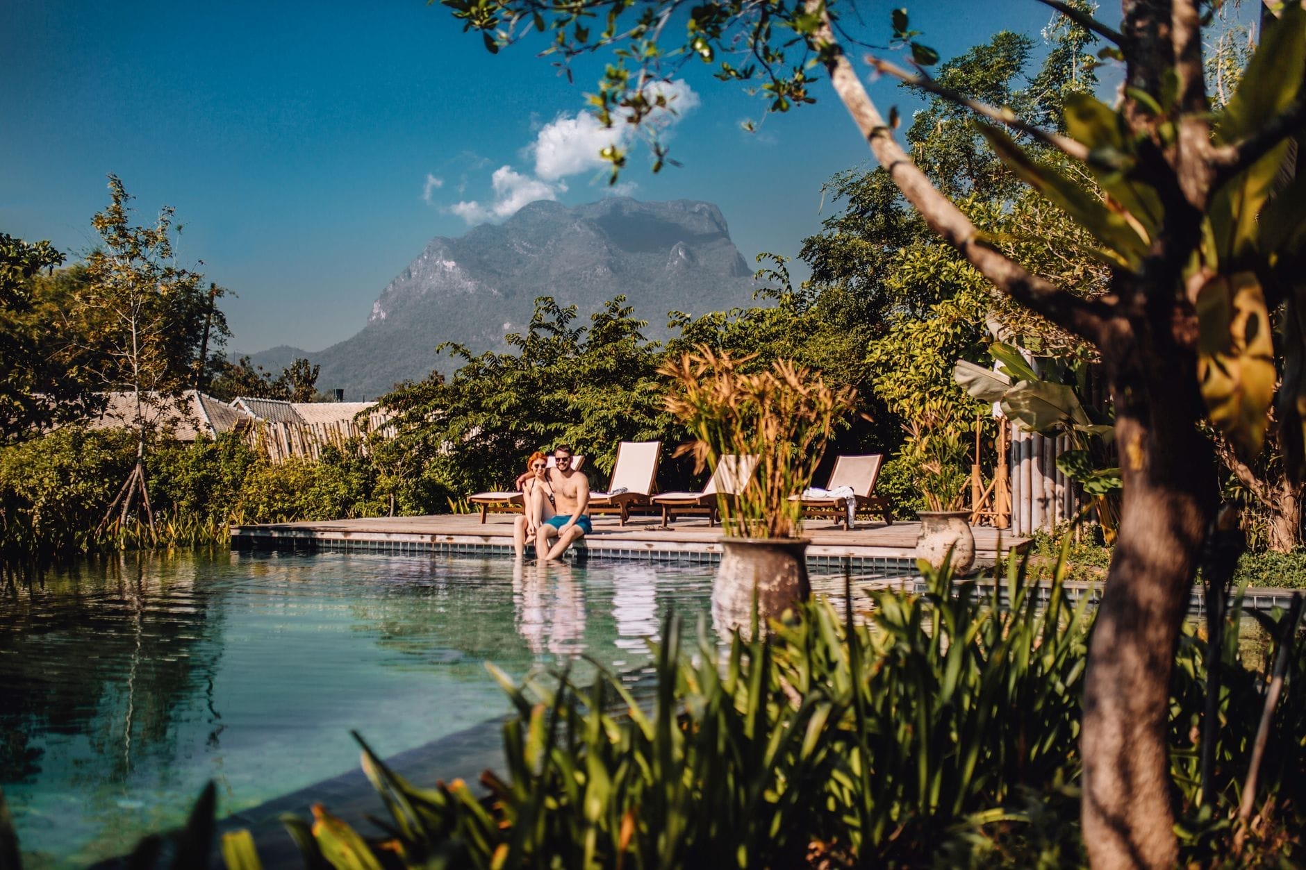 Una pareja se relaja junto a una piscina infinita con vistas a exuberante vegetación y montañas en un resort de lujo en Chiang Mai, con tumbonas cerca.