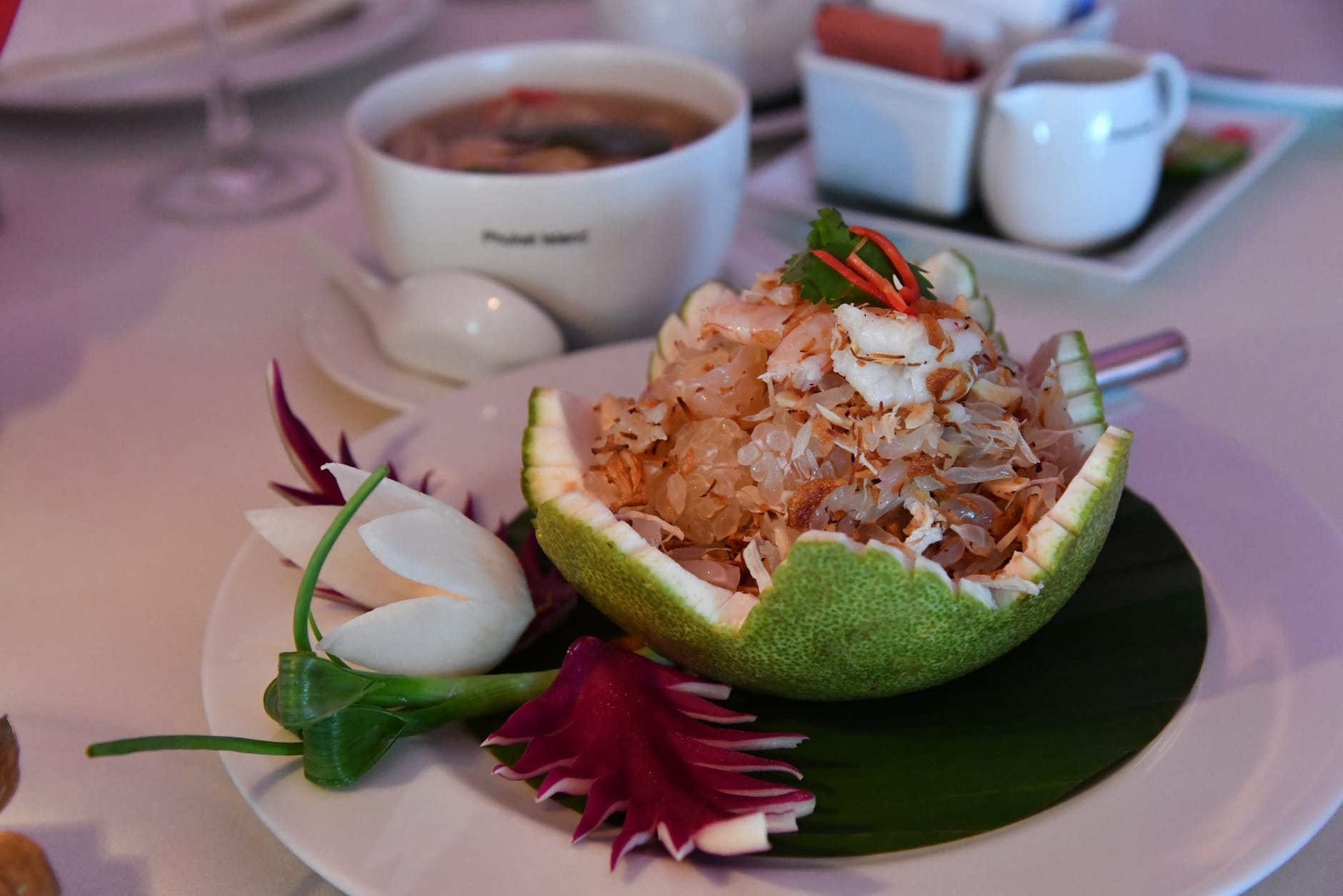 Elegante plato tailandés con comida finamente preparada servida en un pomelo tallado, con adornos artísticos de verduras, sobre una mesa de restaurante.
