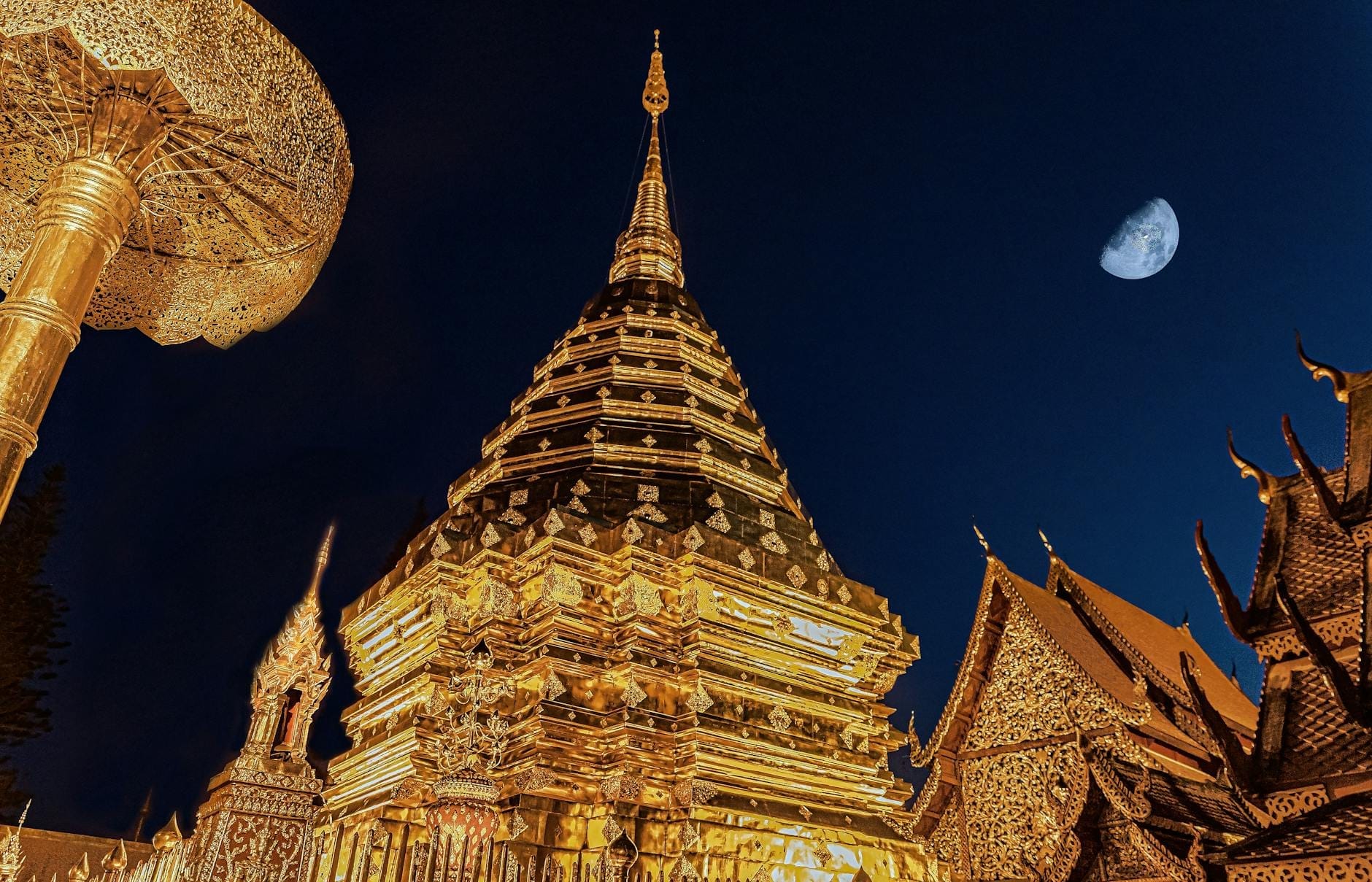 Estupa dorada del Wat Phra That Doi Suthep iluminada de noche con la luna en un cielo azul oscuro, mostrando la atmósfera exclusiva del templo.