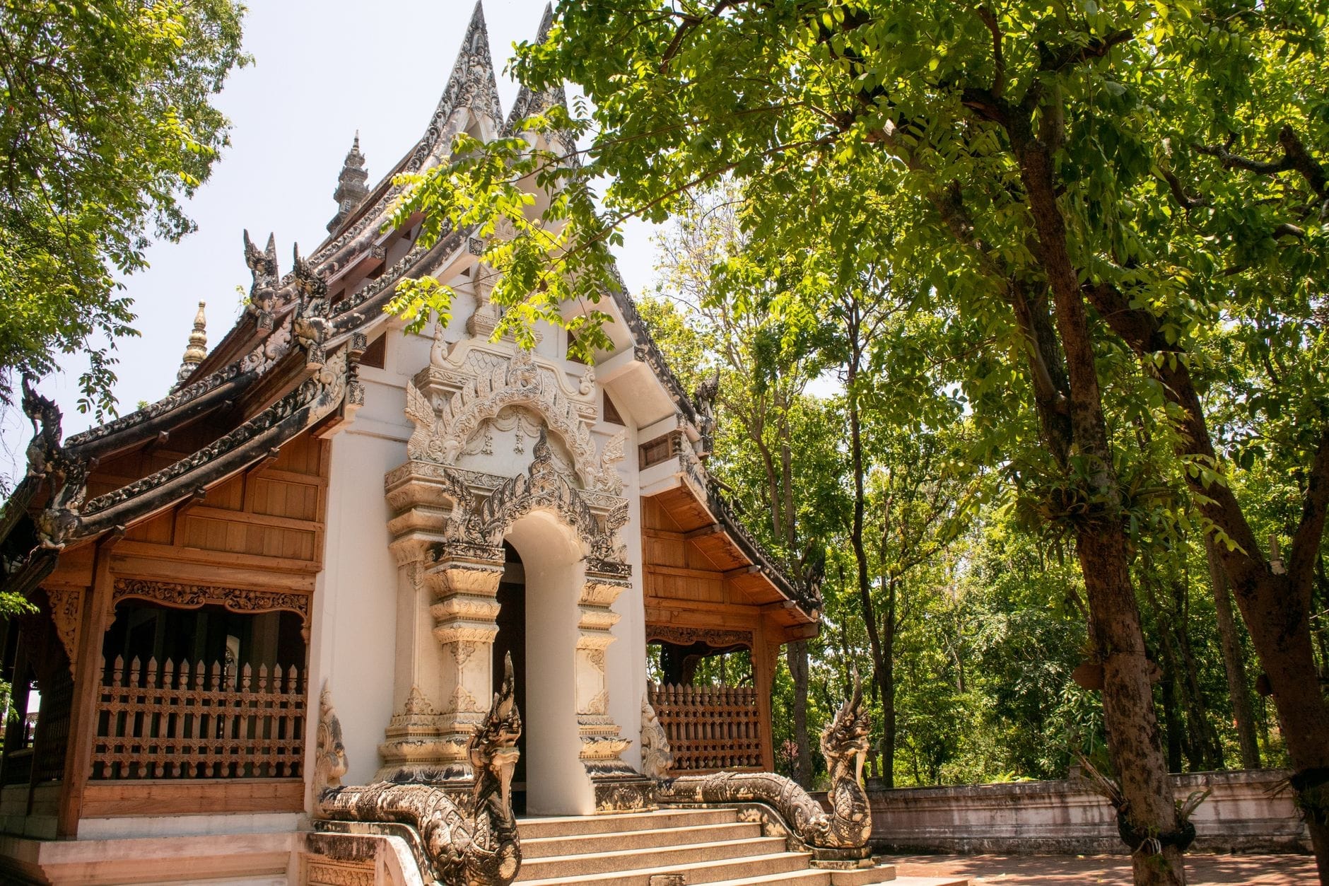 Edificio de templo de estilo Lanna tradicional con intrincadas tallas en madera y fachada blanca, rodeado de frondosos árboles verdes en Chiang Mai.