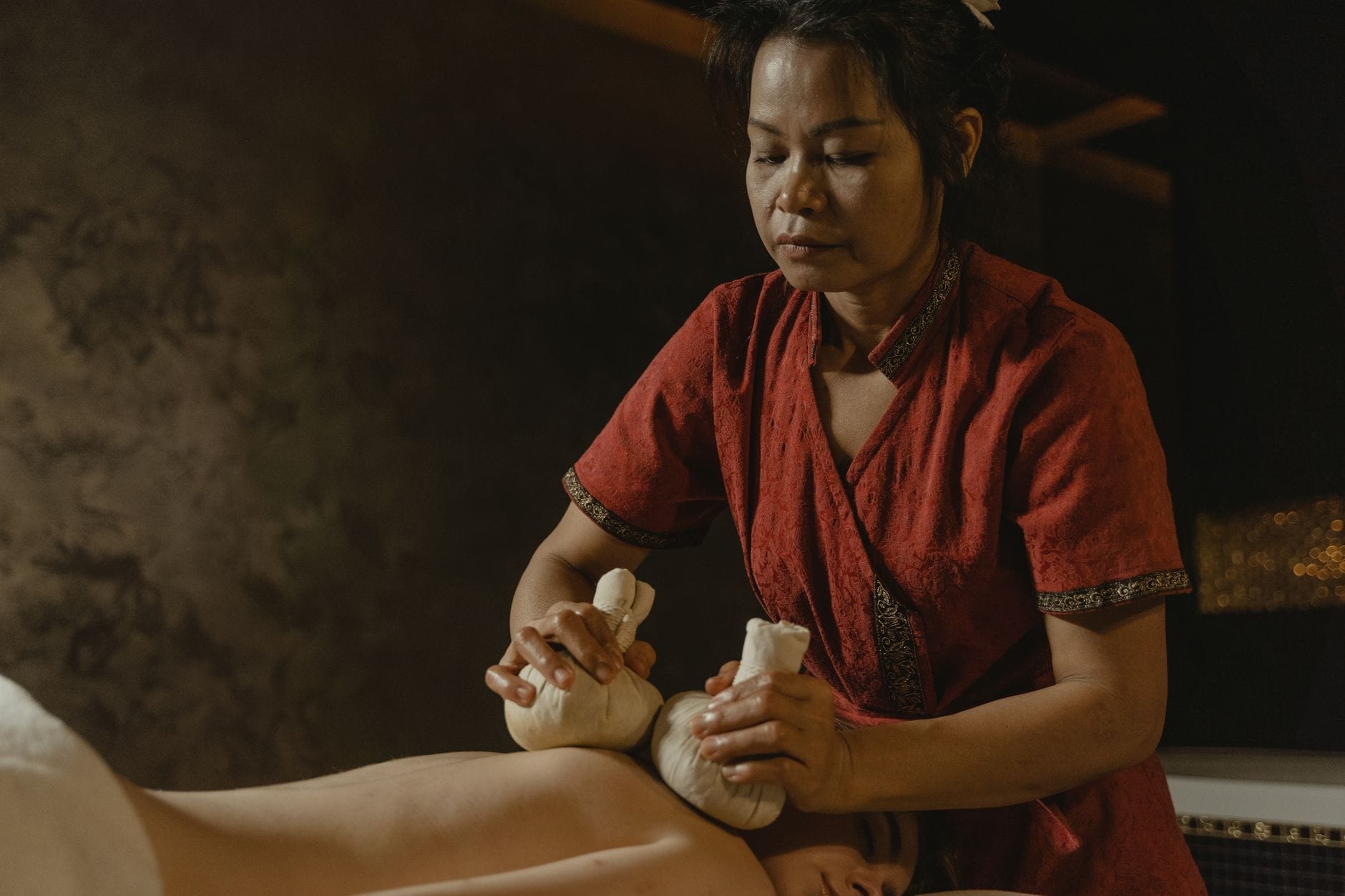 Una mujer vestida con traje tradicional realiza un masaje con compresas de hierbas en un spa con iluminación suave, evocando las tradiciones de bienestar Thai.
