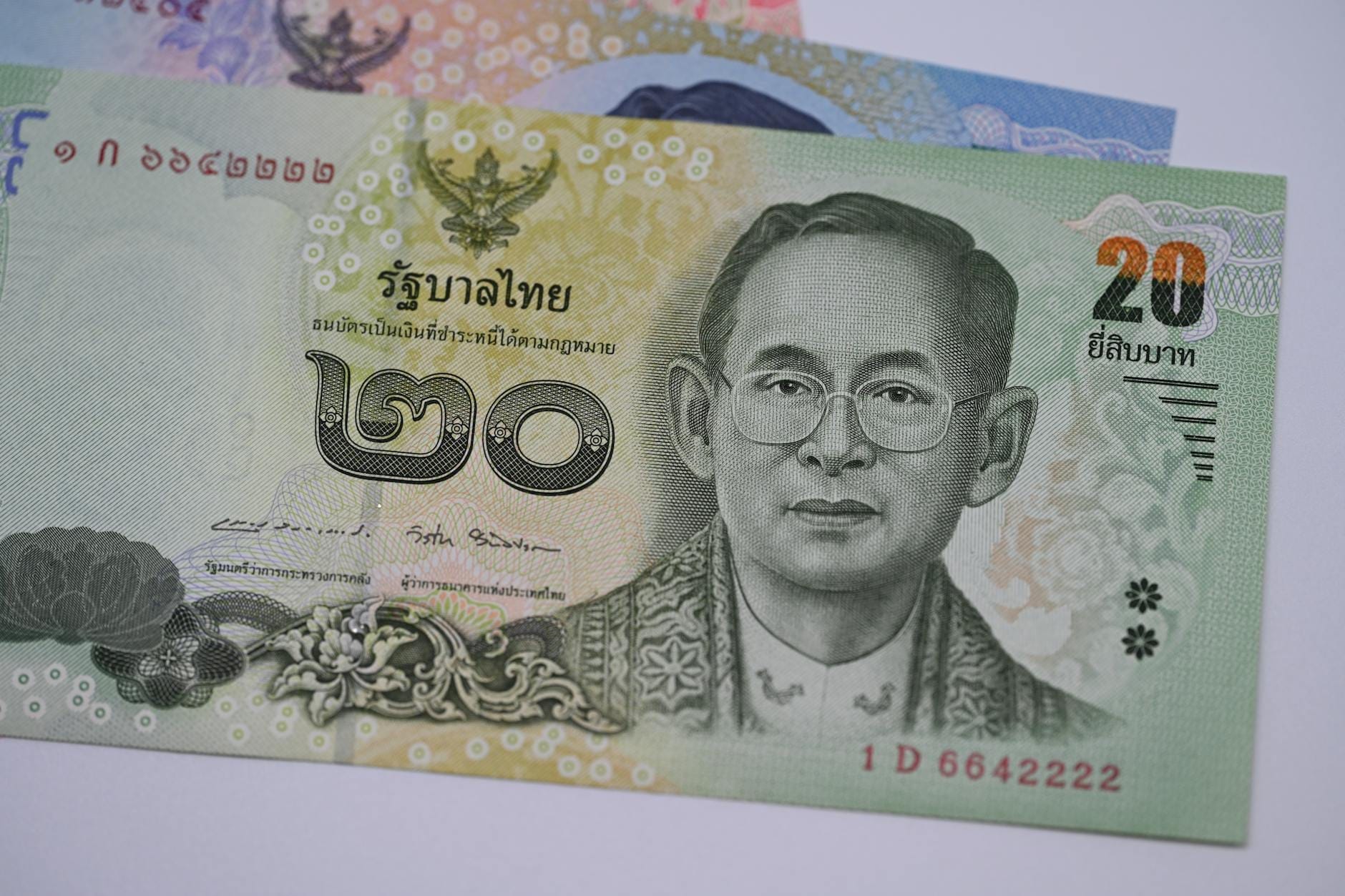 Primer plano de un billete de 20 baht tailandés con el retrato de un rey tailandés, con otros billetes de baht ligeramente visibles al fondo.