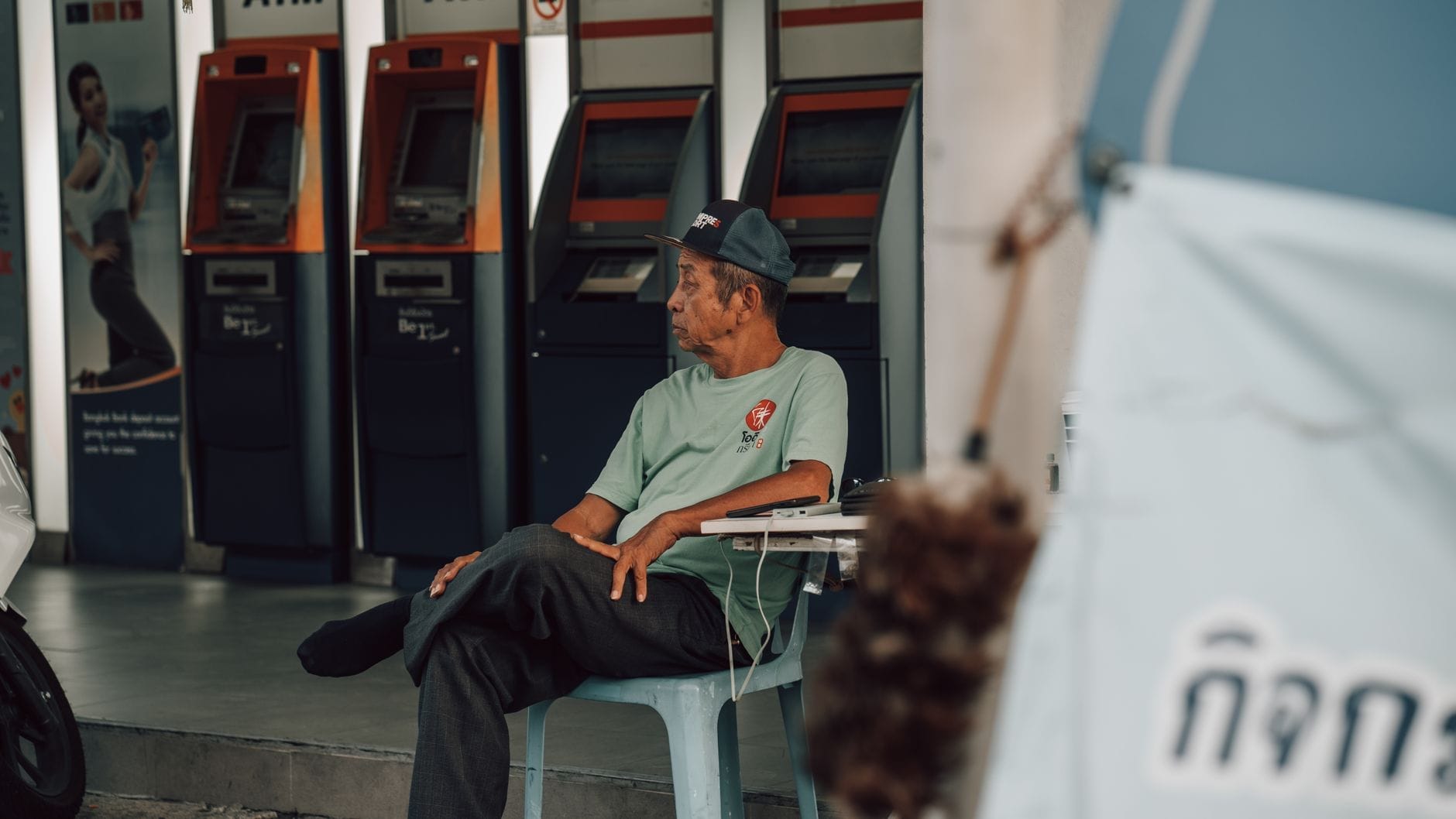 Fila de cajeros automáticos modernos con escritura tailandesa encima, hombre sentado cerca, escena claramente del sudeste asiático típica del Chiang Mai urbano.