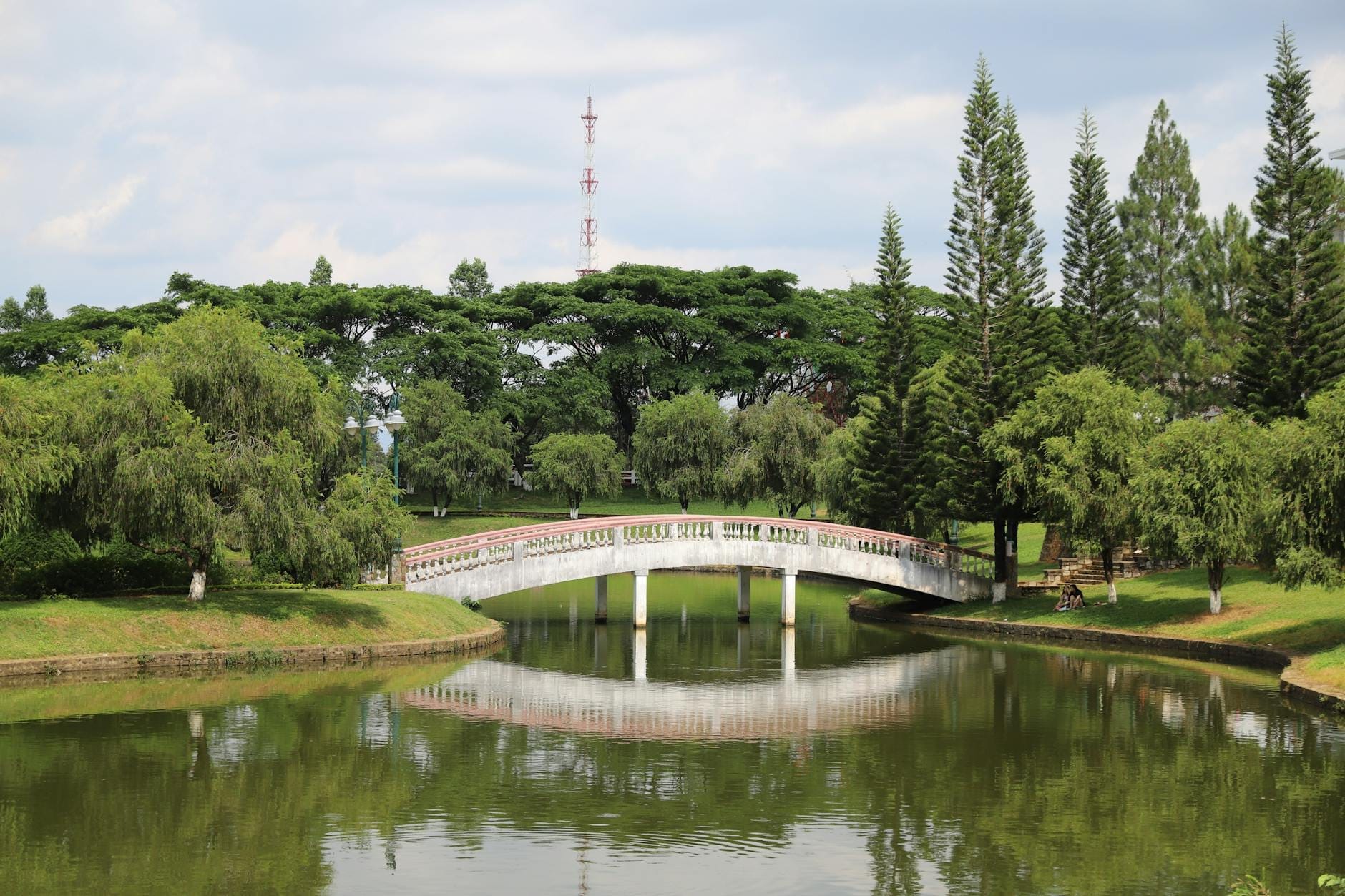 Một công viên yên bình ở Chiang Mai với cây cầu vòm màu trắng bắc qua hồ nước, được bao quanh bởi cây cối và cây xanh tươi tốt, lý tưởng cho một chuyến đi bộ trong thành phố.