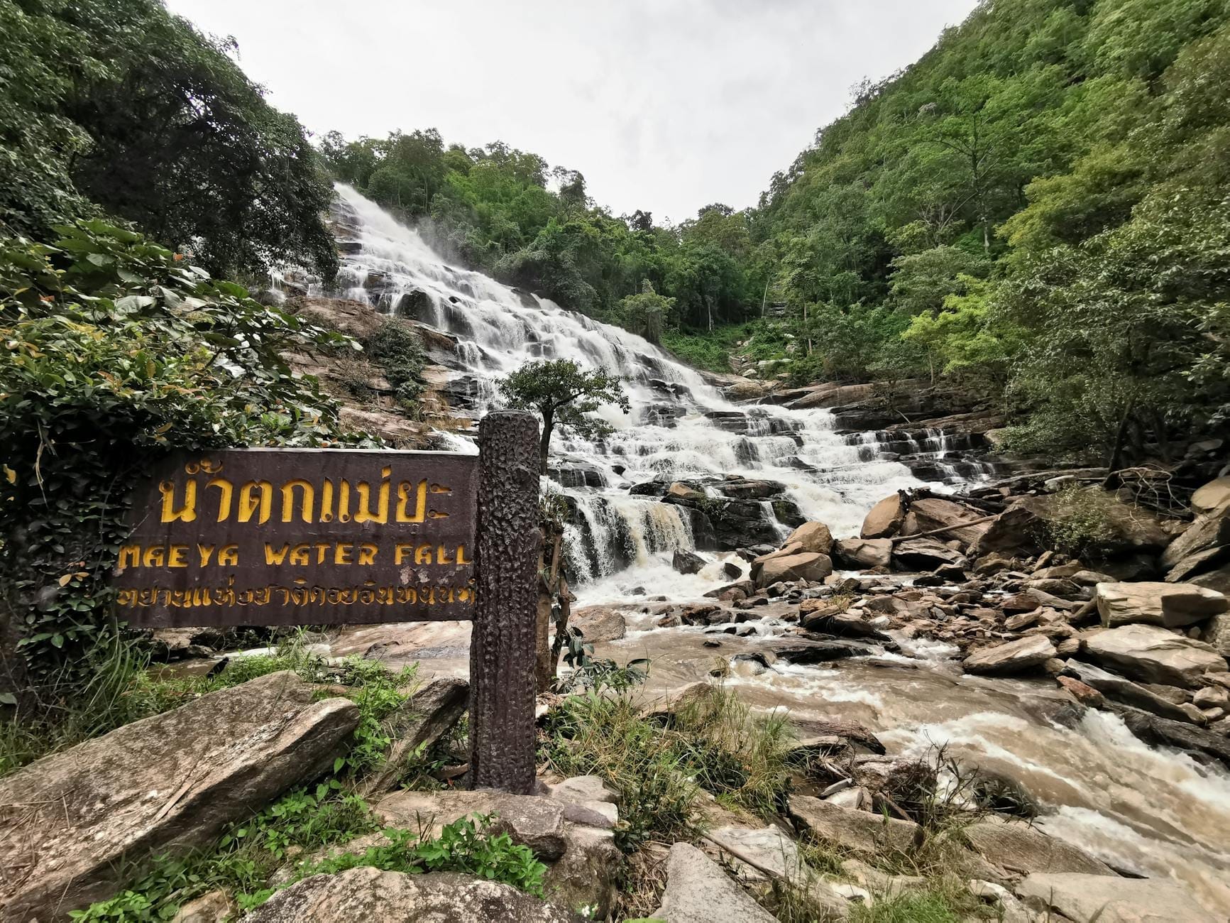Thác nước rộng đổ xuống sườn dốc đầy đá và rừng cây với biển chỉ dẫn Mae Ya Waterfall gần Chiang Mai ở phía trước.