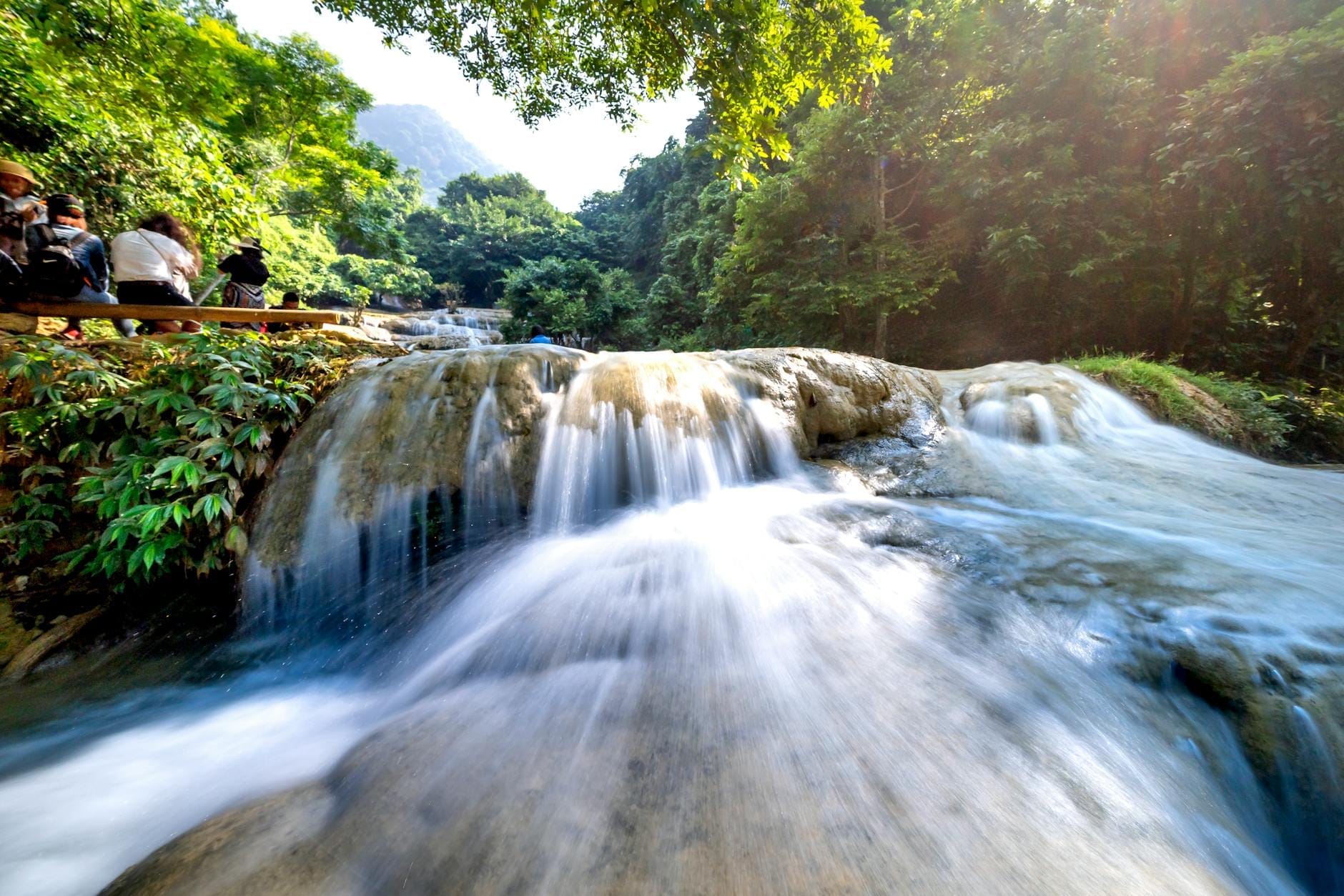 Một thác nước nhiều tầng tuyệt đẹp đổ xuống những tảng đá nhẵn bóng được bao quanh bởi khu rừng xanh tươi tốt, với du khách ngồi bên cạnh thưởng thức cảnh quan thiên nhiên.