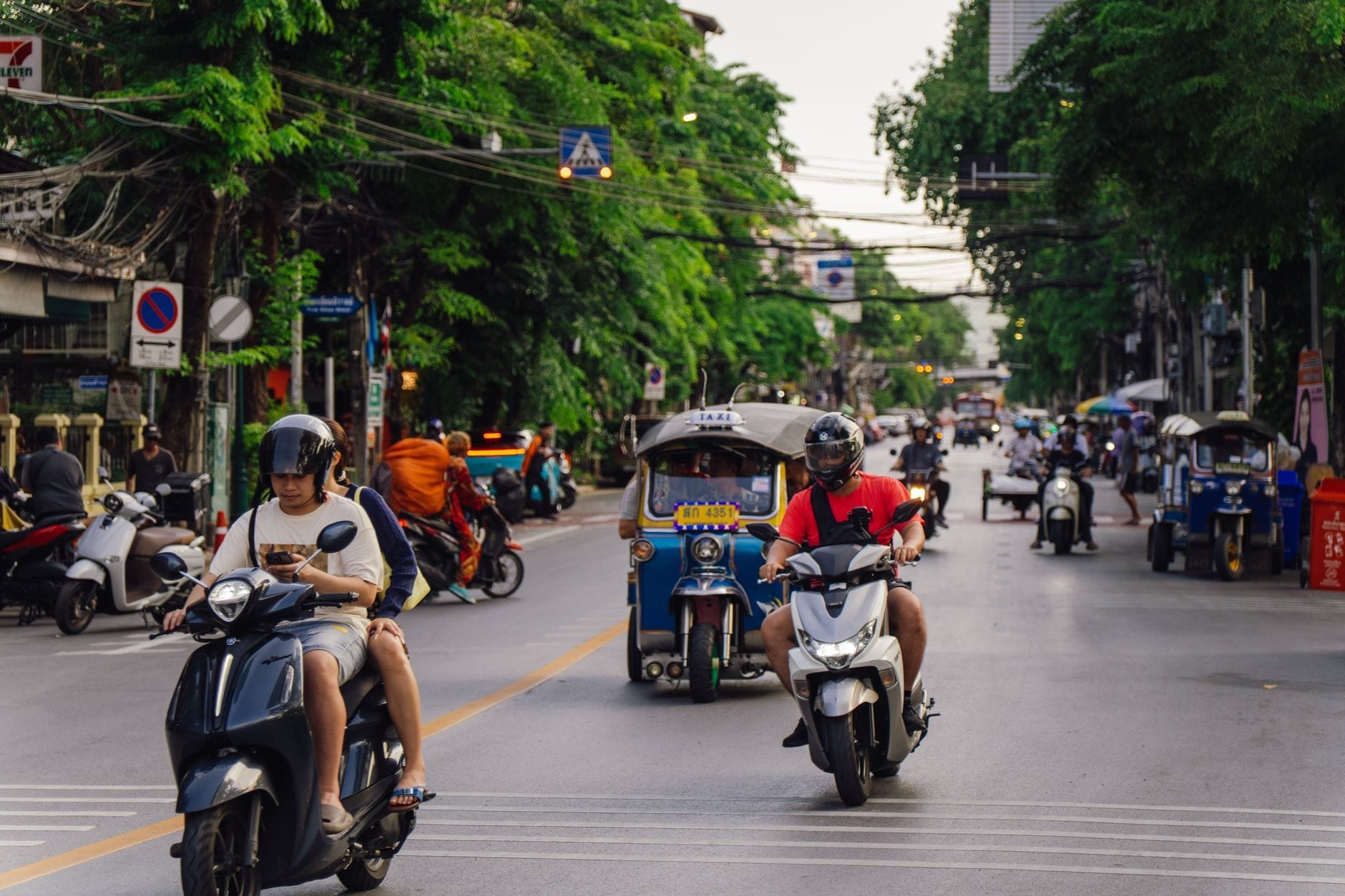 Concurrida calle de Chiang Mai con varias personas en motocicletas, un tuk tuk y edificios urbanos al fondo.