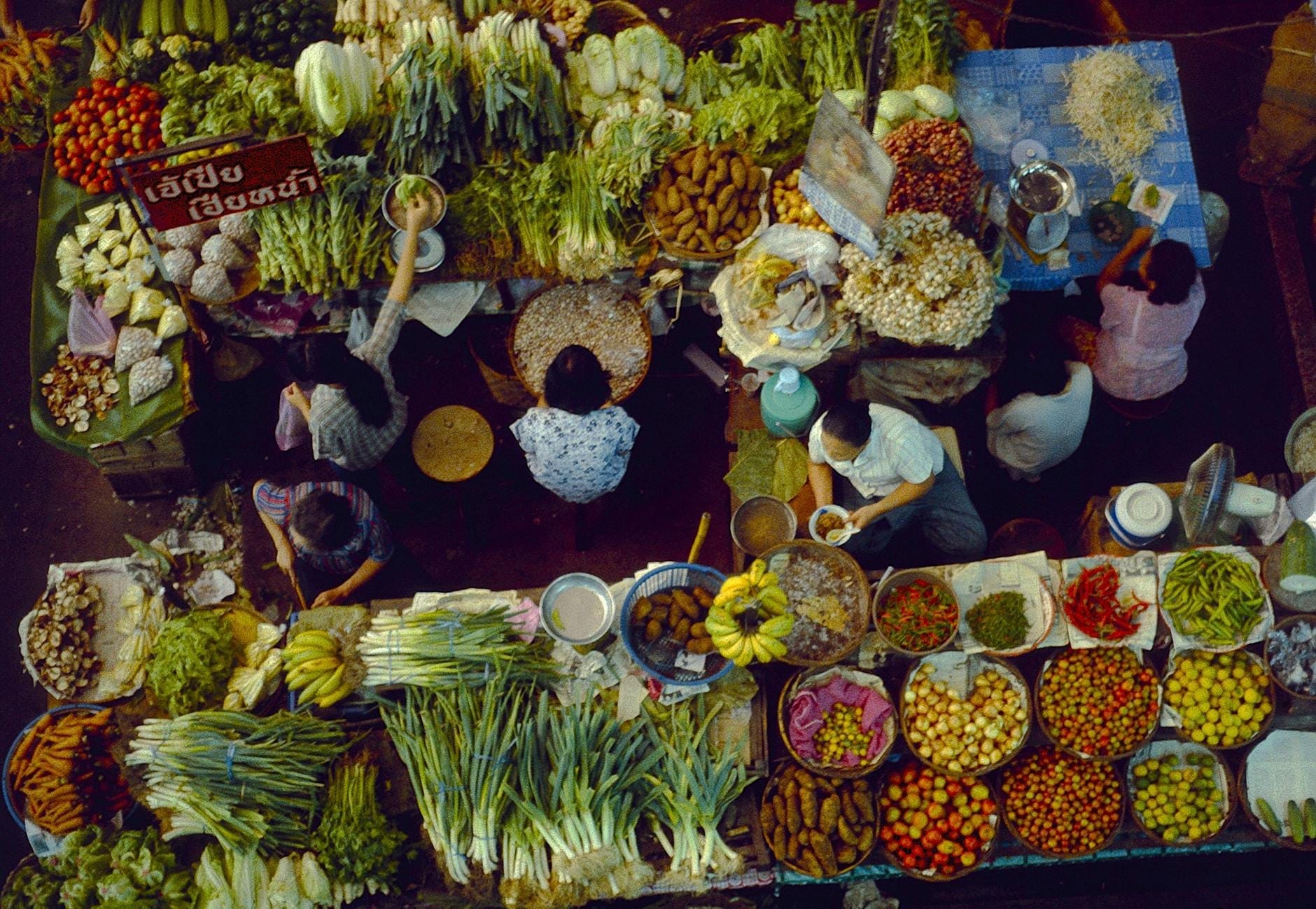 Vista aérea de un animado mercado de Chiang Mai con vendedores ofreciendo verduras frescas, frutas y hierbas en puestos coloridos, lleno de gente comprando.