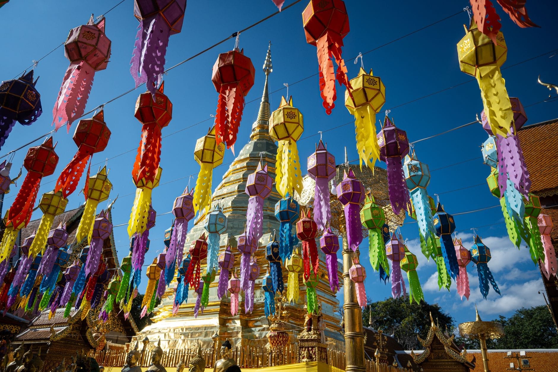 Coloridas linternas de papel colgando en el cielo con un templo dorado de Chiang Mai y cielo azul de fondo, capturando la atmósfera festiva de Tailandia.