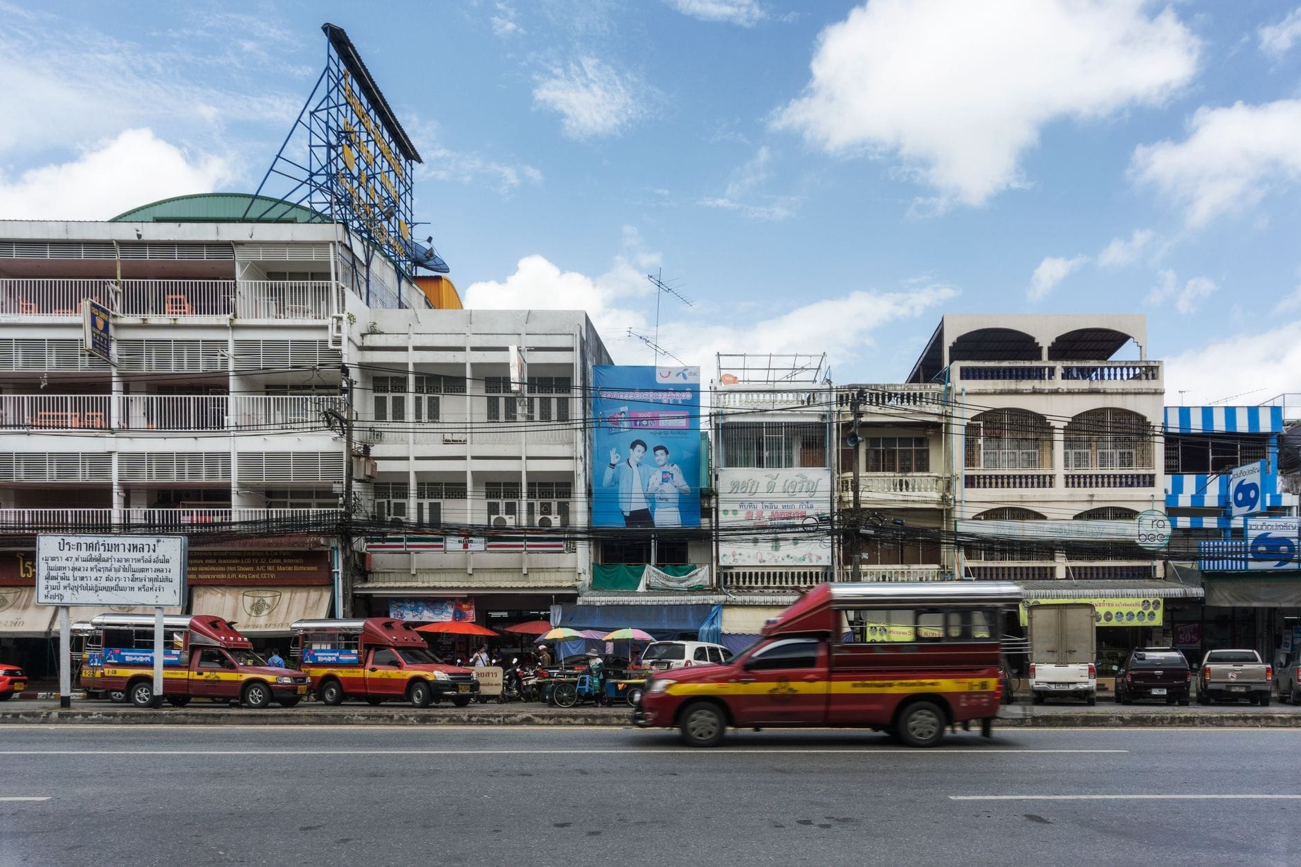 Cảnh đường phố ở Chiang Mai với những chiếc songthaew đỏ cổ điển (xe taxi chung), cửa hàng và các tòa nhà địa phương dưới bầu trời xanh, nổi bật các lựa chọn phương tiện giao thông địa phương.