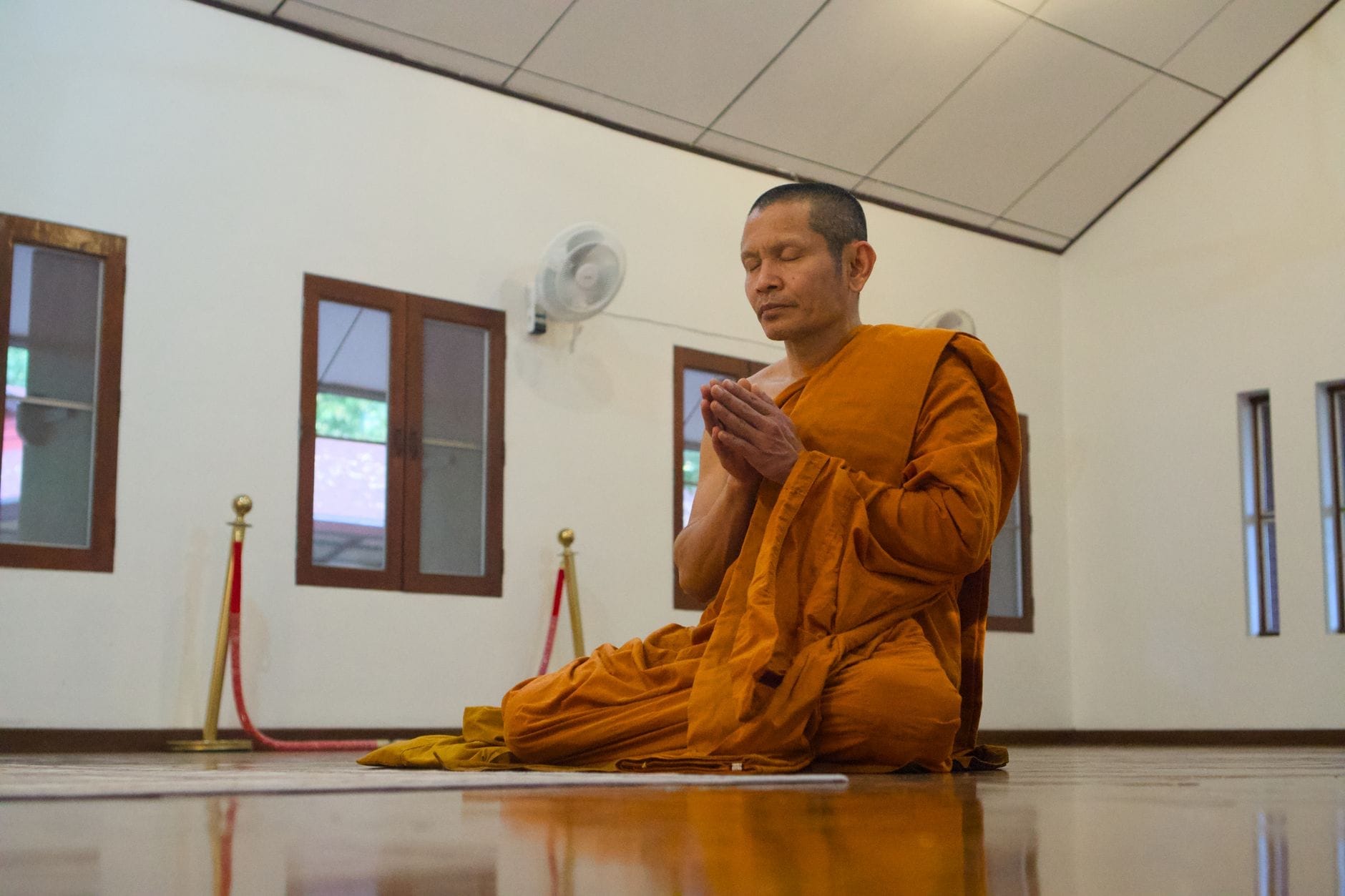 Một nhà sư Phật giáo mặc áo cà sa màu cam đang thiền định trong một căn phòng chùa yên tĩnh với sàn gỗ và trang trí đơn giản tại Chiang Mai.