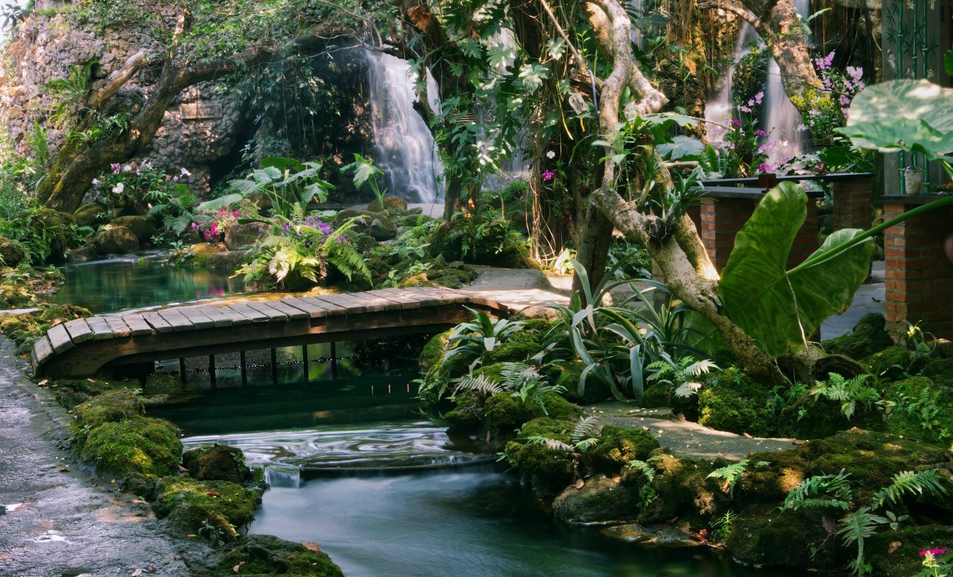 Jardín de spa con exuberante vegetación, pequeño puente de madera sobre un arroyo, rodeado de plantas tropicales, flores y cascadas en un ambiente tranquilo.