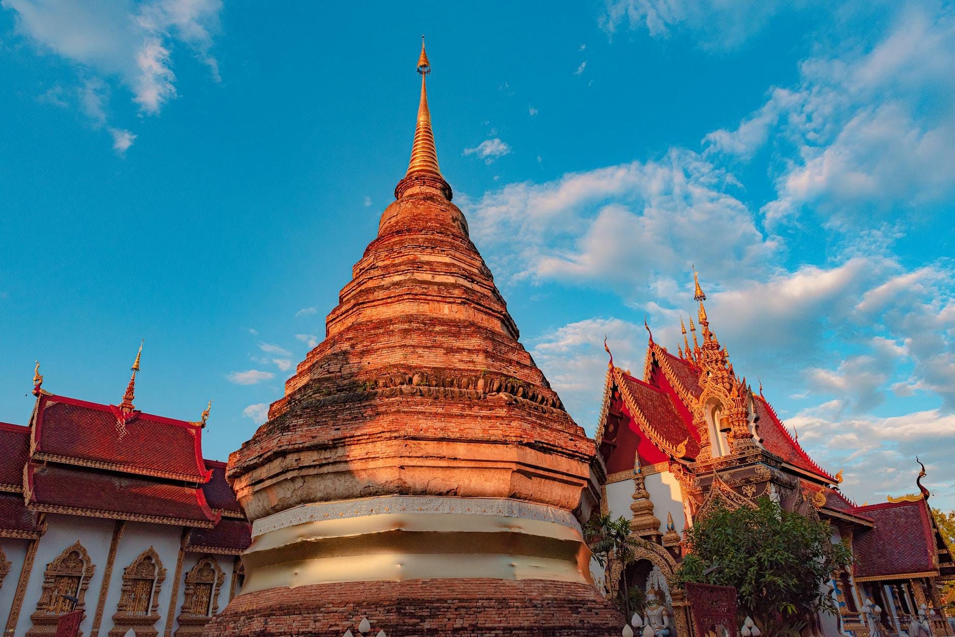 Quần thể đền chùa Chiang Mai ngập nắng với một bảo tháp vàng và những mái nhà chạm trổ tinh xảo dưới bầu trời xanh với những đám mây rải rác.
