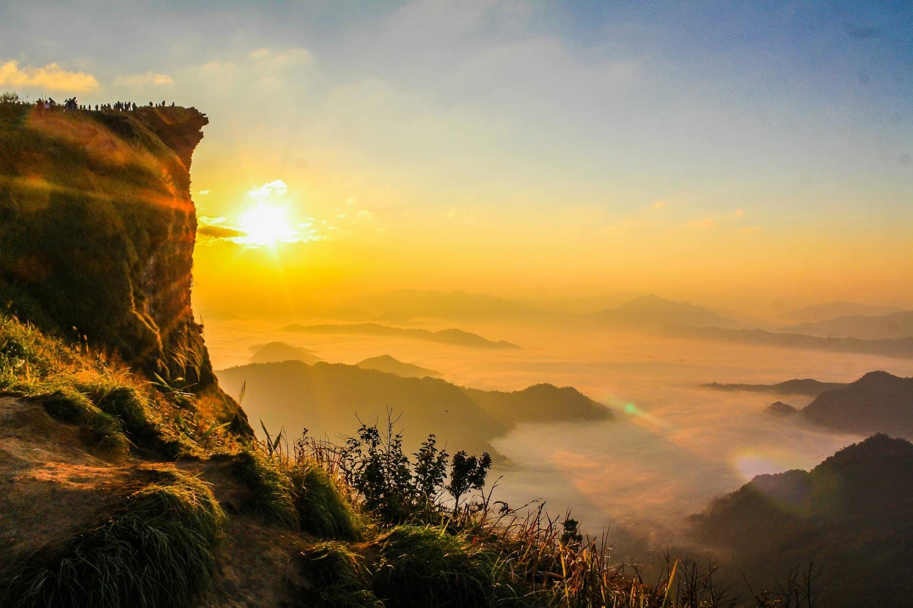 Amanecer dorado sobre montañas y valles brumosos en el norte de Tailandia con personas reunidas en un acantilado contemplando la escena.