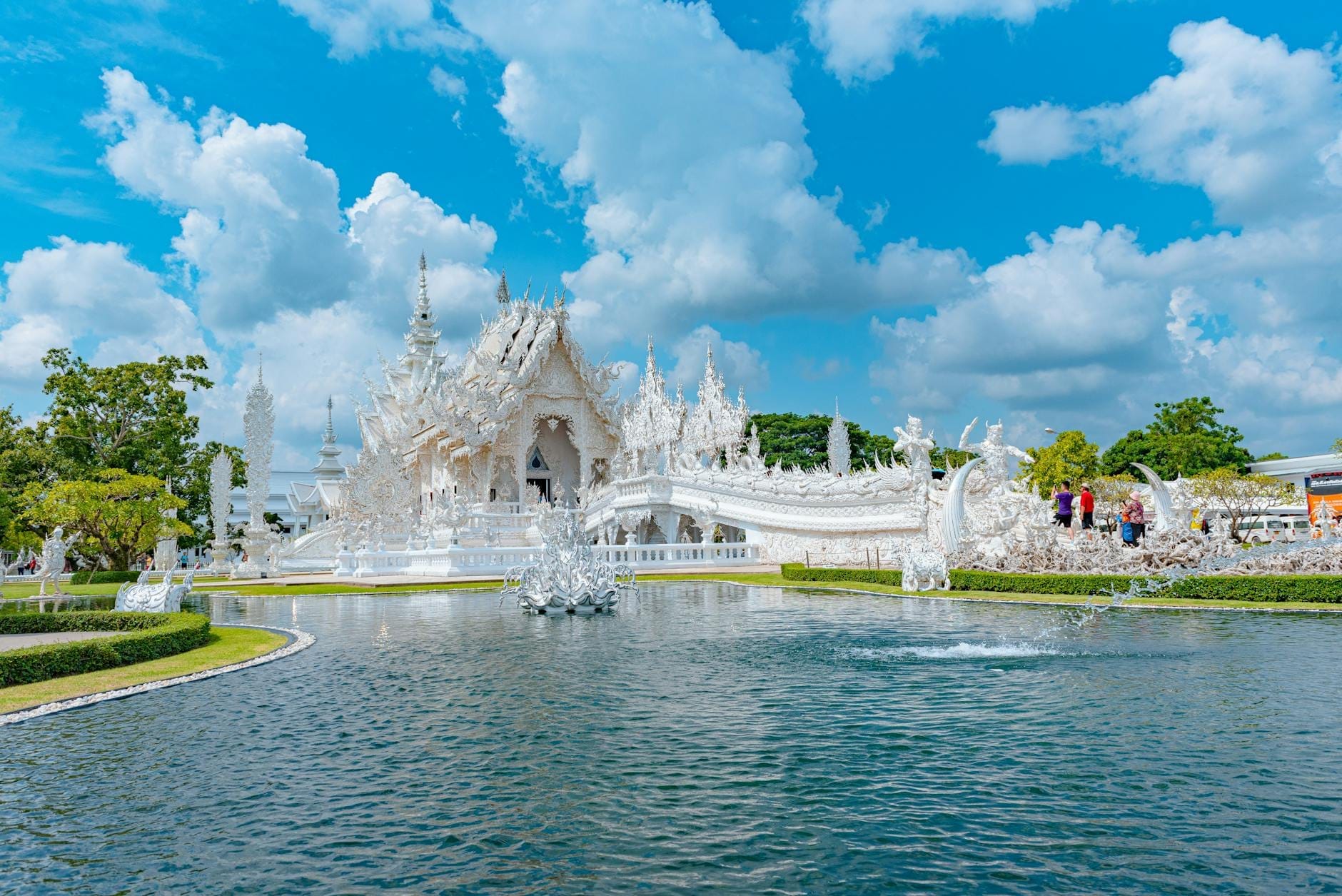 El Templo Blanco (Wat Rong Khun) en Chiang Rai, Tailandia, con su ornamentada arquitectura blanca y su puente reflejados en un estanque en un día soleado.