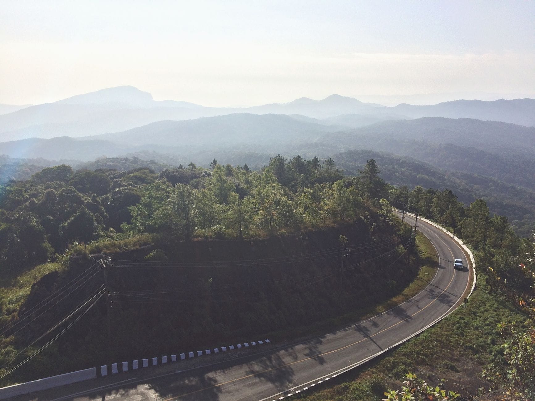Carretera de montaña serpenteante con un coche, rodeada de vegetación exuberante y colinas neblinosas en la ruta entre Chiang Mai y Chiang Rai al amanecer.