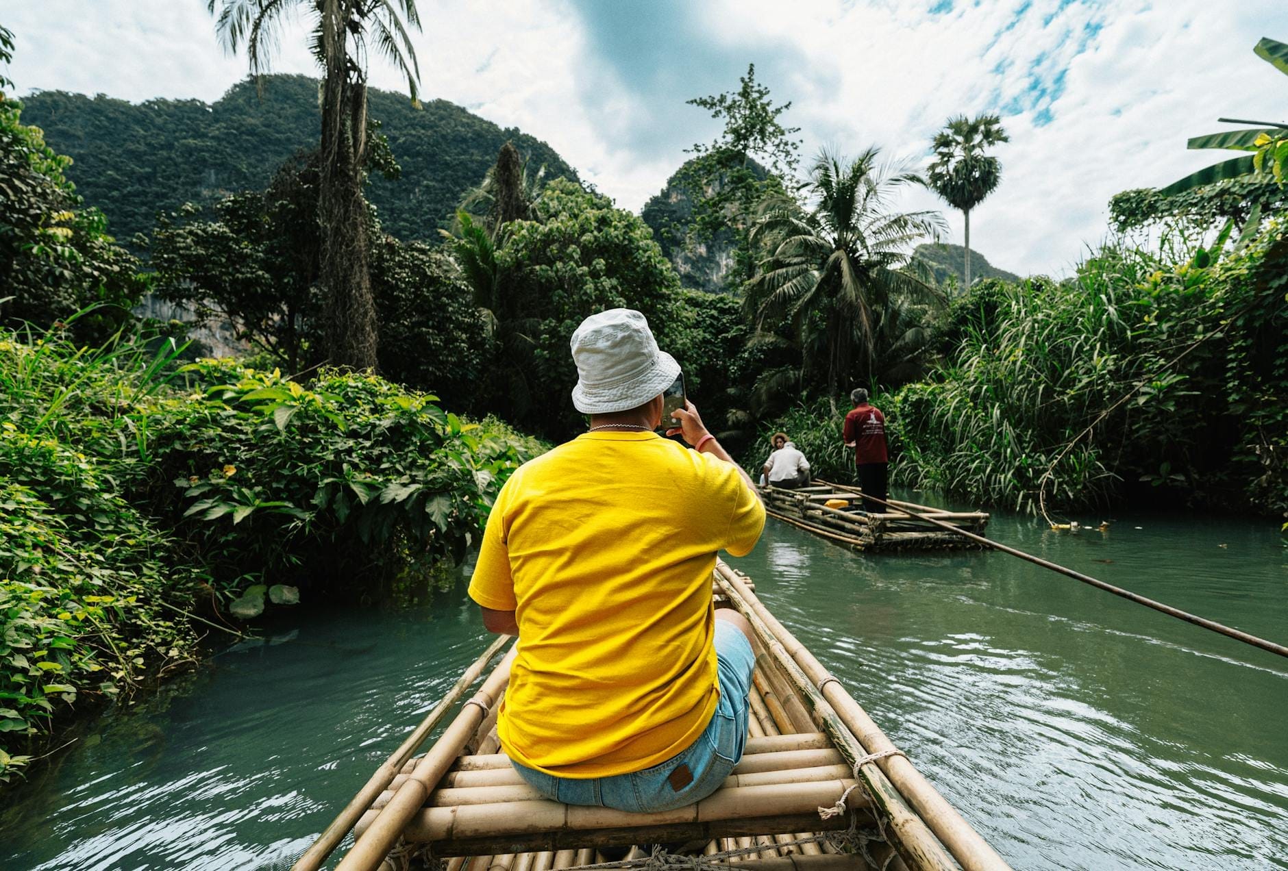 Du khách trên bè tre đang di chuyển dọc theo dòng sông trong rừng rậm cùng hướng dẫn viên địa phương, bao quanh là thảm thực vật xanh tươi và những ngọn núi xa xa.