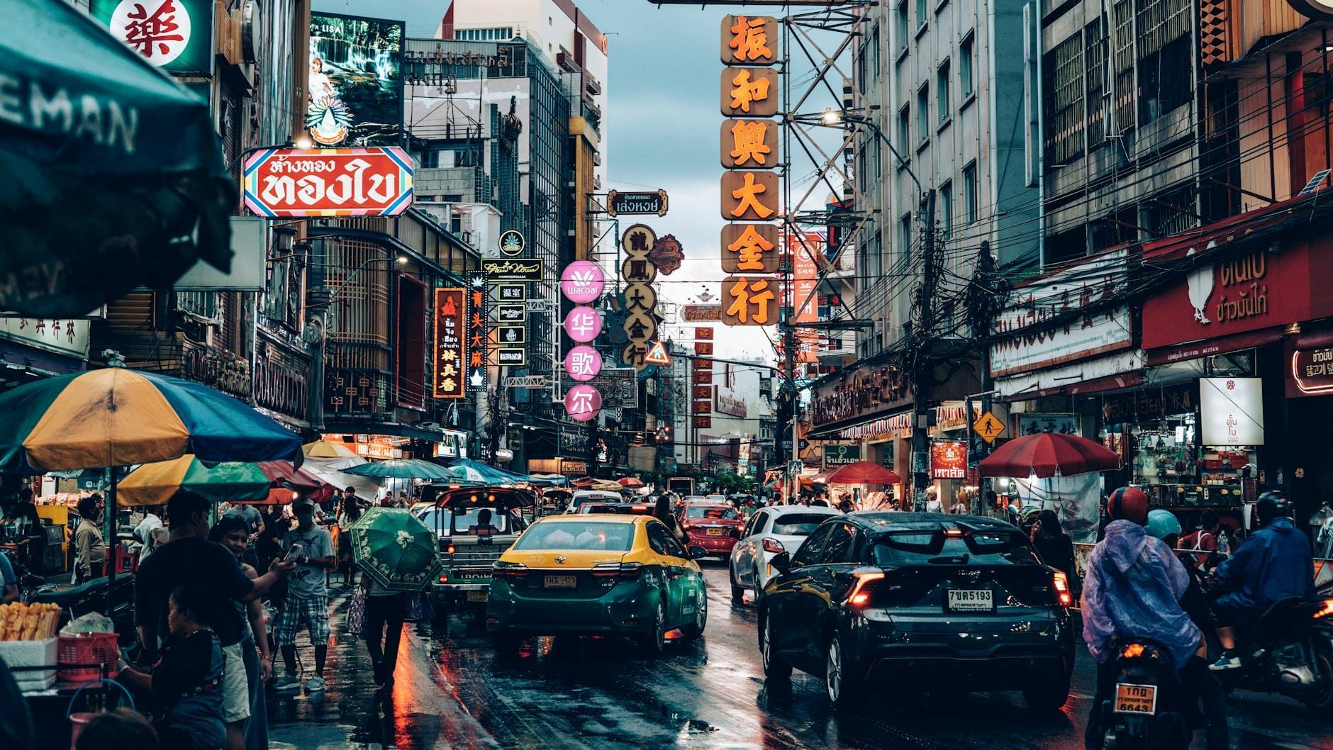 Concurrida escena callejera en el barrio chino de Bangkok al atardecer, con letreros de neón, taxis, motocicletas, vendedores ambulantes y personas bajo coloridos paraguas.