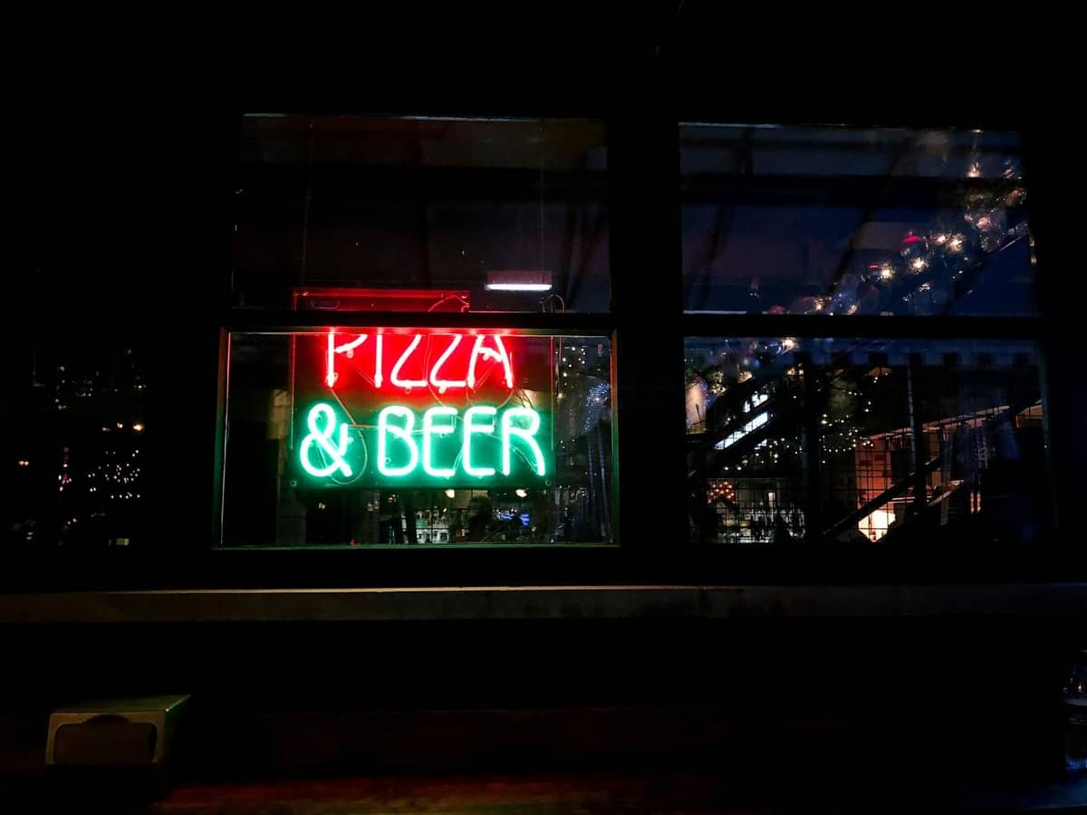 Neonschild in einem Fenster bei Nacht mit der Aufschrift Pizza & Beer und Spiegelungen der Stadtlichter, das eine lebhafte urbane Abendszene andeutet.