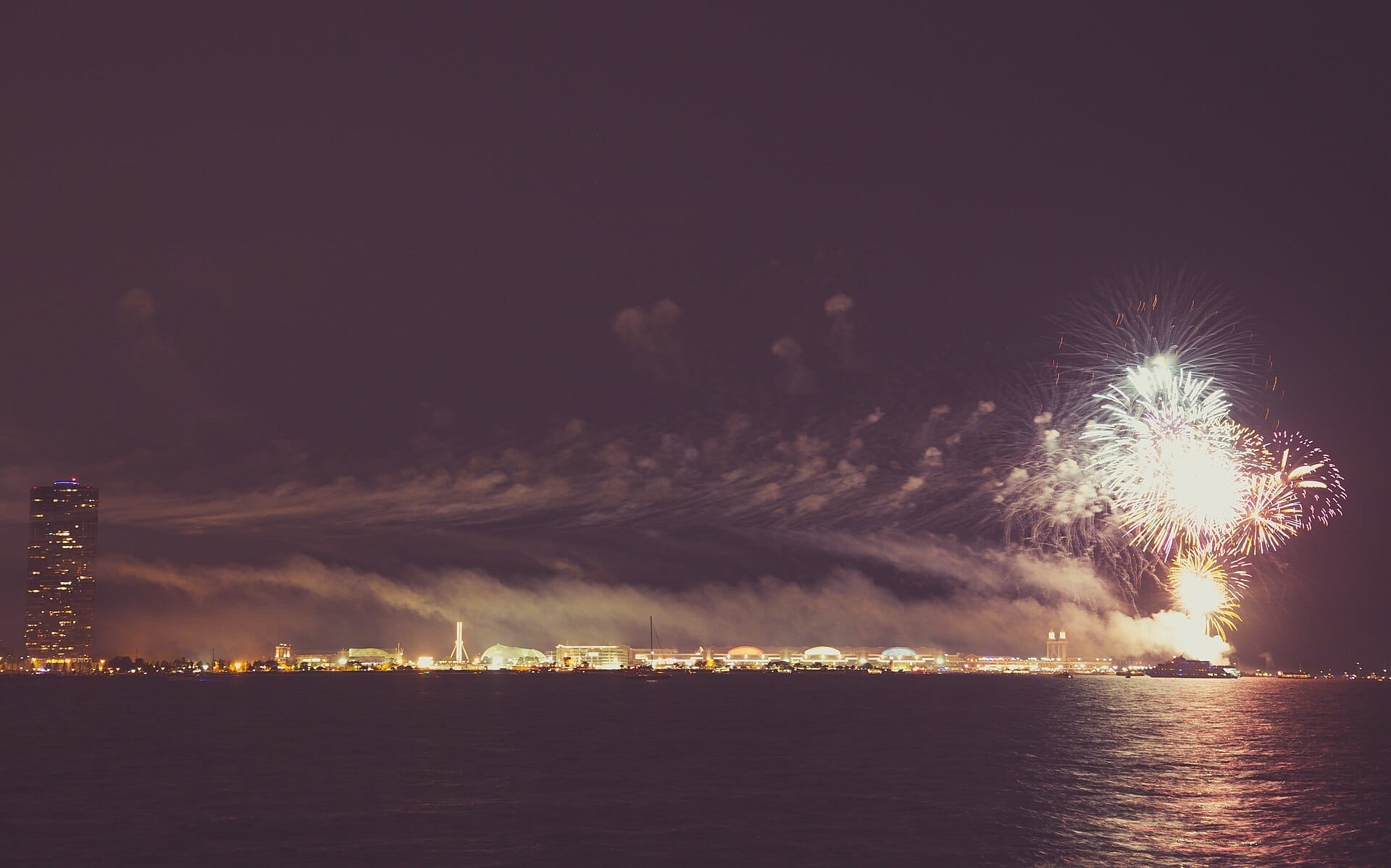 Fogos de artifício coloridos explodem sobre o Navy Pier e o lago Michigan à noite, com um arranha-céu e as luzes da cidade visíveis ao longo da margem.