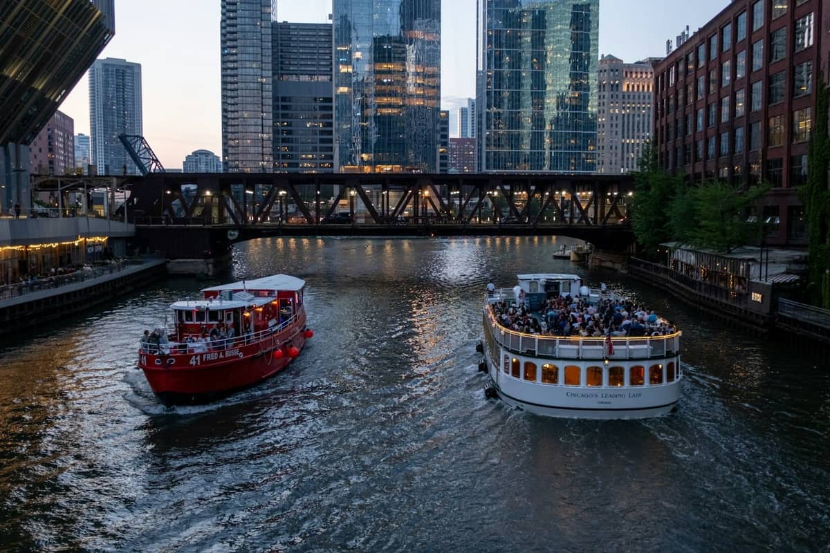 Dos barcos turísticos llenos de personas navegan por el río Chicago al atardecer con rascacielos iluminados y un puente a la vista.