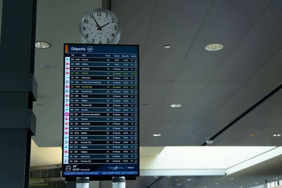 Tablica odlotów na lotnisku pokazująca wiele lotów i destynacji linii lotniczych, z zegarem powyżej i nowoczesnym sufitem terminalu w tle.