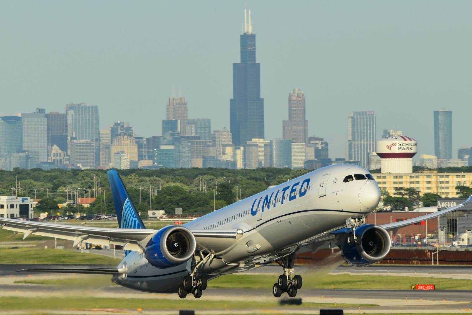 Um avião da United decolando em um aeroporto de Chicago com o skyline da cidade e a Willis Tower visíveis ao fundo em um dia ensolarado.