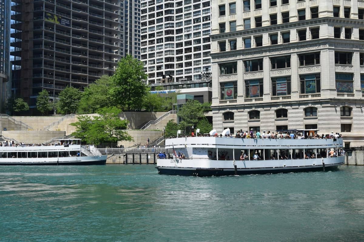 Two architecture tour boats docked by the Chicago Riverwalk, with city buildings in the background on a sunny day.