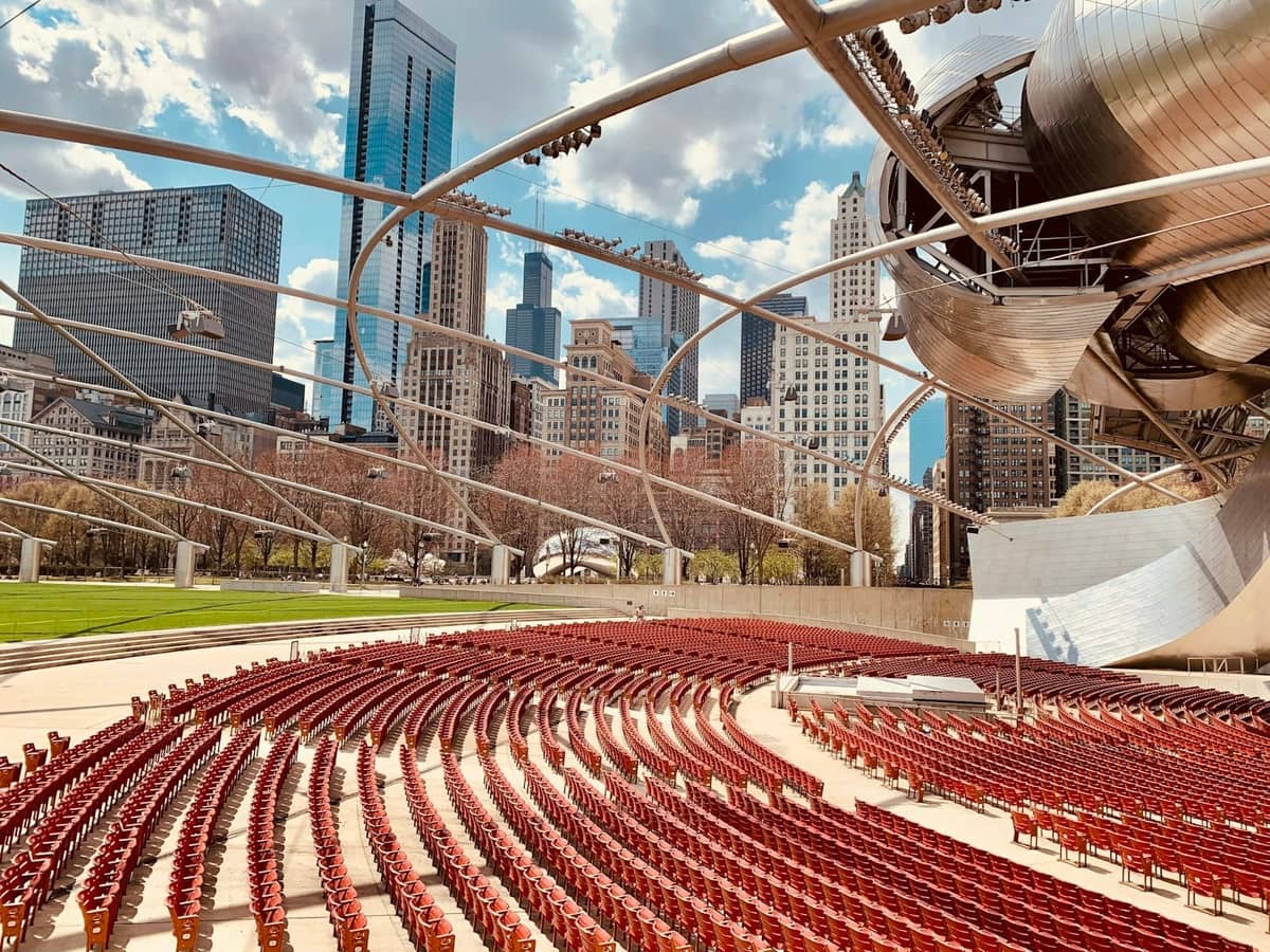 Leerer Außensitzbereich des Jay Pritzker Pavilion im Millennium Park mit der Skyline von Chicago im Hintergrund, unter einem geschwungenen Stahlgitterwerk.