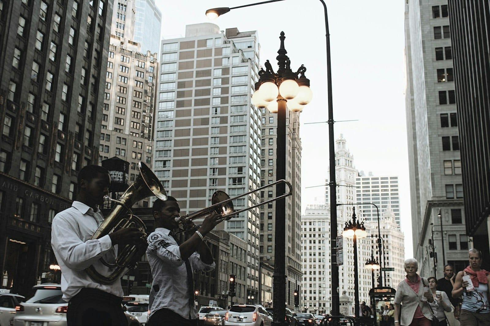 Músicos de rua tocam instrumentos de sopro no centro de Chicago, rodeados por altos edifícios, postes de luz brilhantes e trânsito urbano ao fundo.