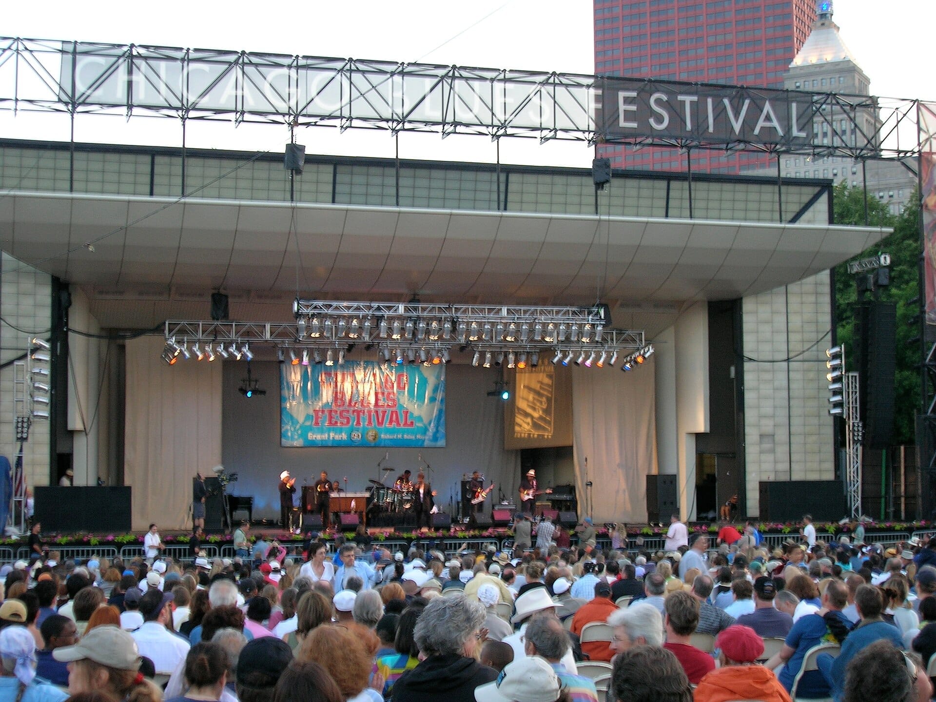 Una grande folla si raduna al Millennium Park per guardare i musicisti esibirsi su un palco all'aperto durante il Chicago Blues Festival sotto un cartello del festival.