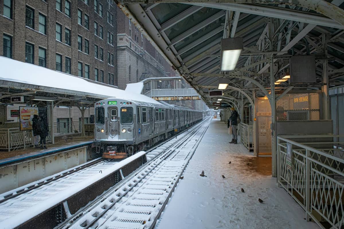 Chicago CTA-Zug, der im Winter an einem verschneiten Hochbahnsteig ankommt, mit wartenden Menschen und umliegenden Stadtgebäuden.