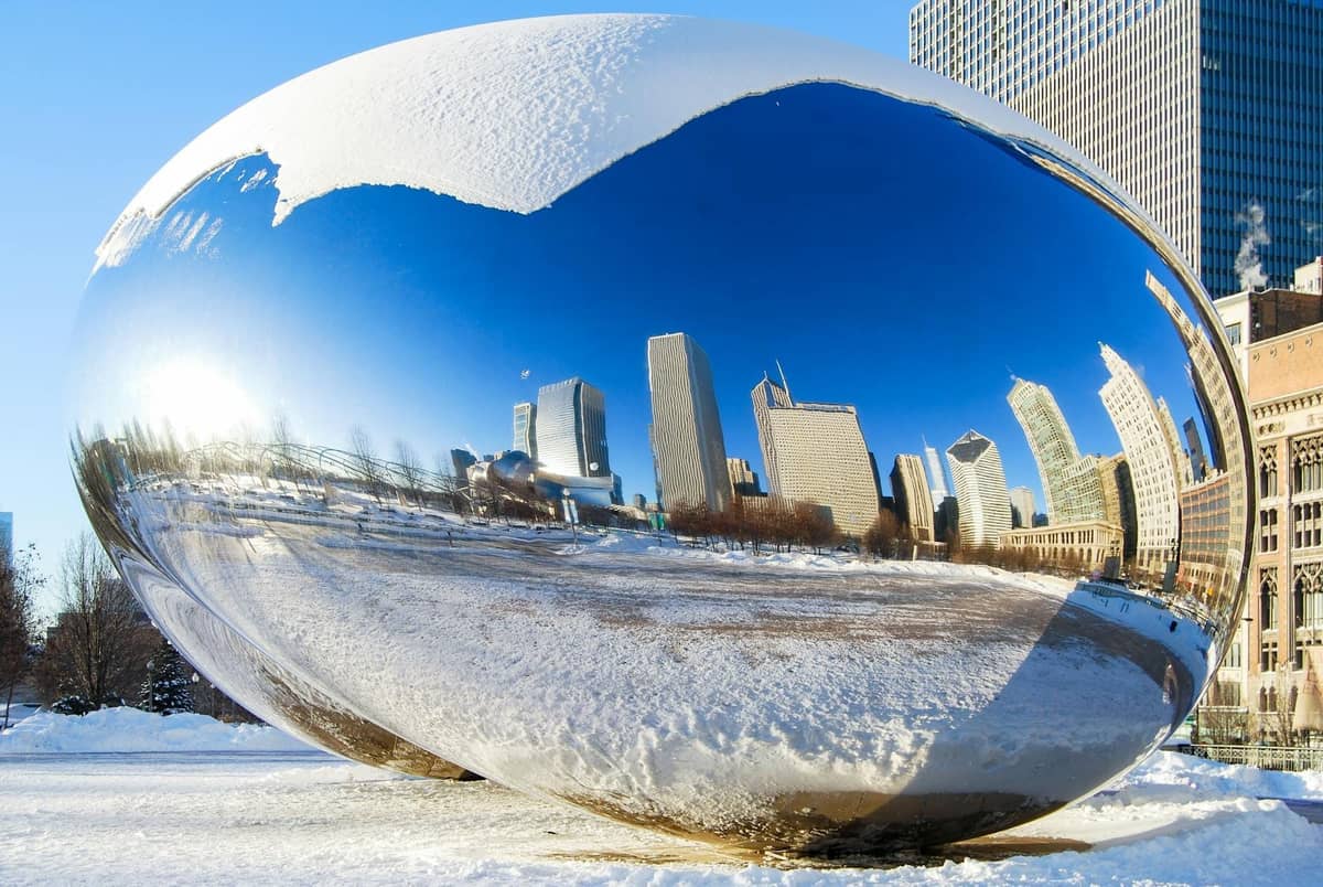 Skulptur Cloud Gate, auch bekannt als 'The Bean', mit Schnee bedeckt und den Wolkenkratzern von Chicago auf ihrer Oberfläche gespiegelt, umgeben von einer Winterlandschaft im Millennium Park.