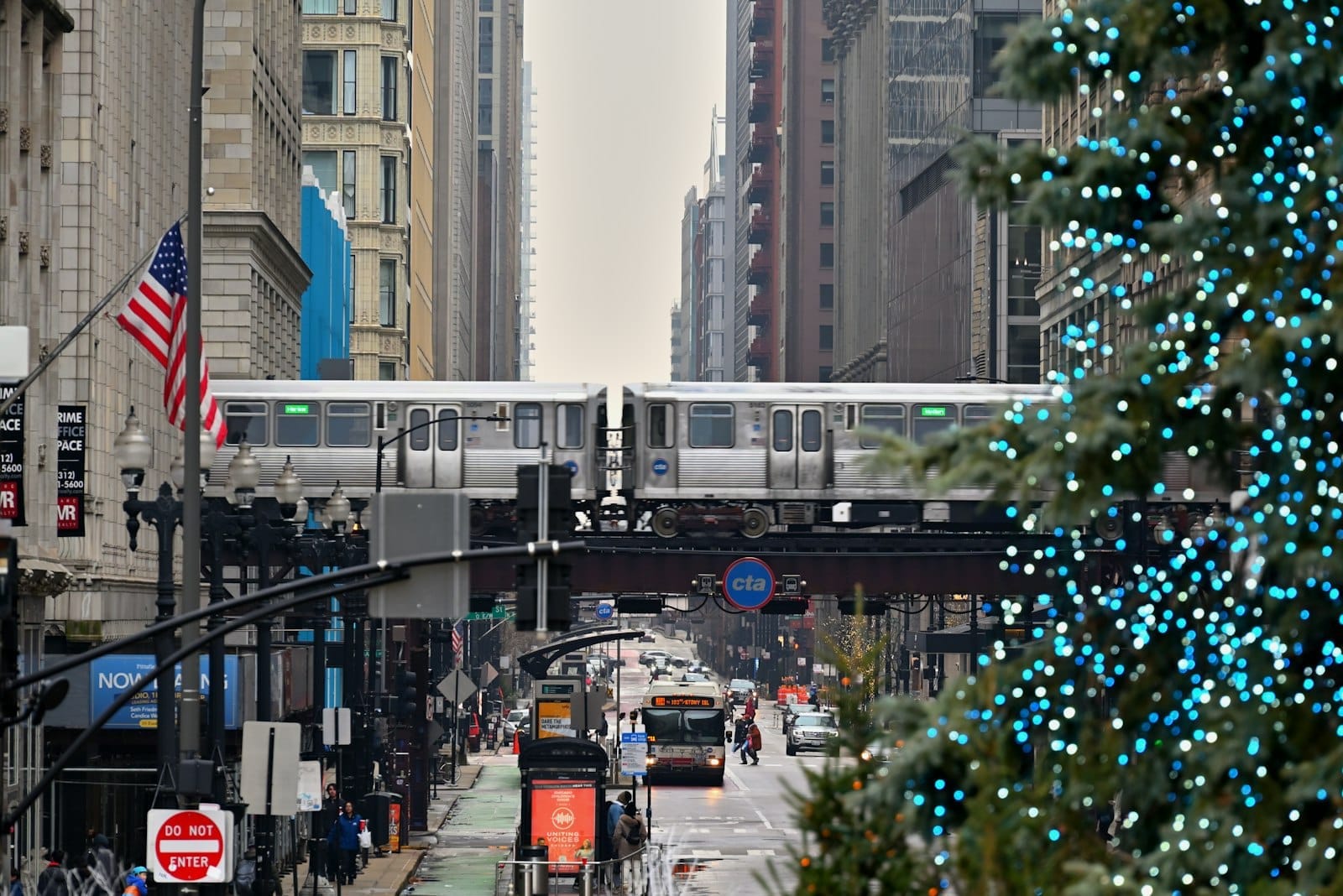 Vista do centro de Chicago com árvore de Natal decorada com luzes azuis, trem elevado passando ao fundo e prédios altos em um dia nublado de inverno.