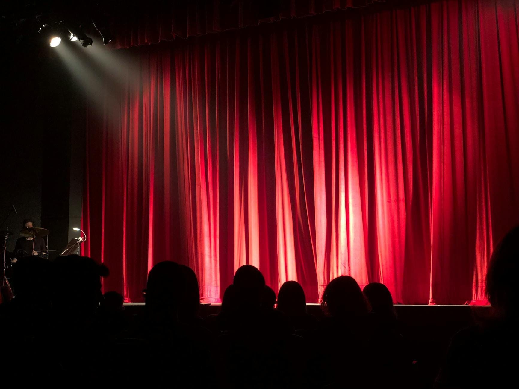 Um teatro com pouca iluminação com cortinas de palco vermelhas fechadas, uma plateia em silhueta em primeiro plano e um músico sentado na bateria à esquerda.