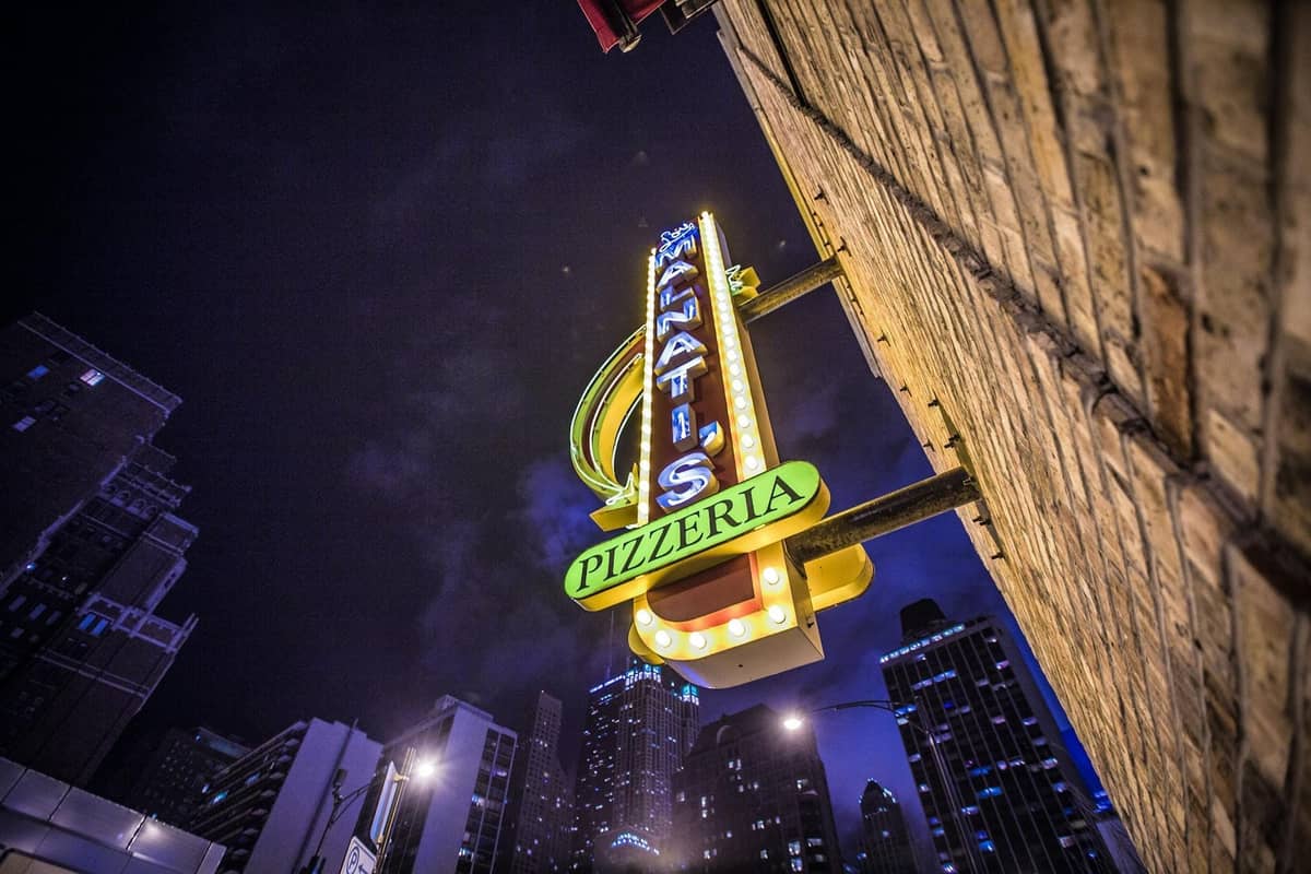 Helles Neonschild einer bekannten Chicago Deep-Dish-Pizzeria, mit der Skyline der Stadt und Hochhäusern, die nachts sichtbar sind.