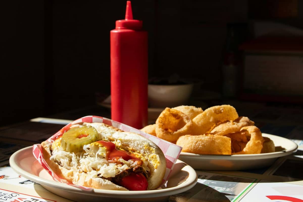 Un clásico hot dog estilo Chicago con pepinillos, tomates, cebollas y mostaza en un panecillo, servido con aros de cebolla y un frasco rojo de condimento.
