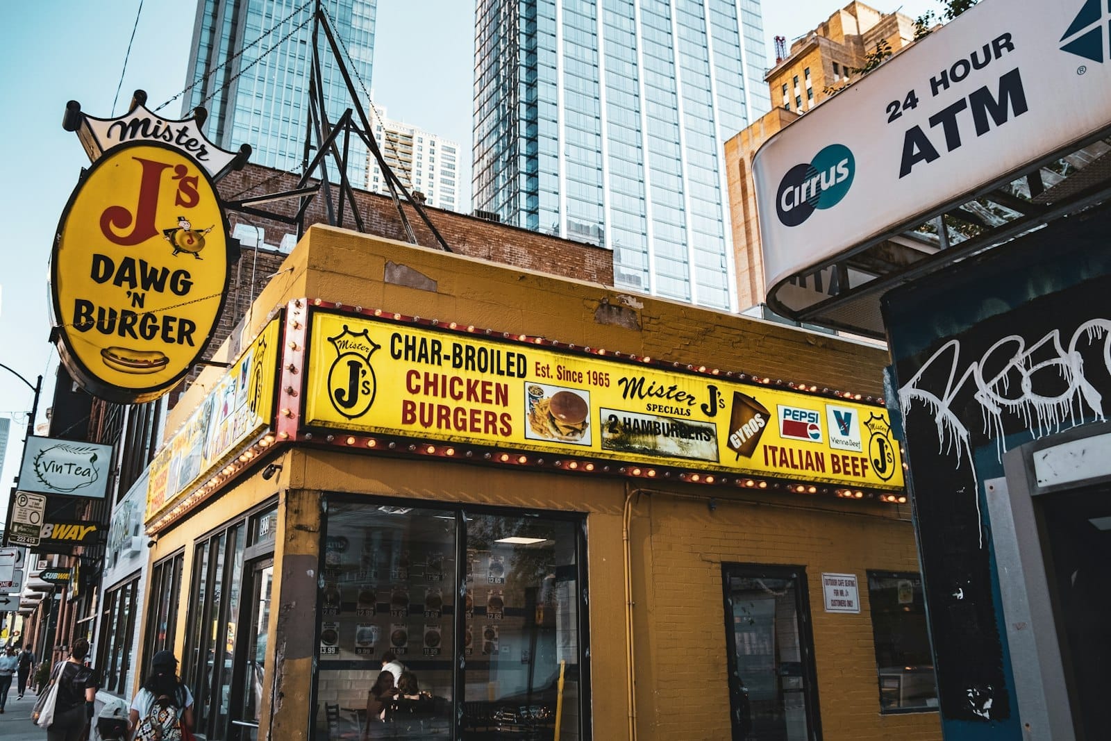 Uma barraca de comida de rua colorida em Chicago com placas para pratos clássicos como Italian beef e hambúrgueres, com arranha-céus ao fundo.